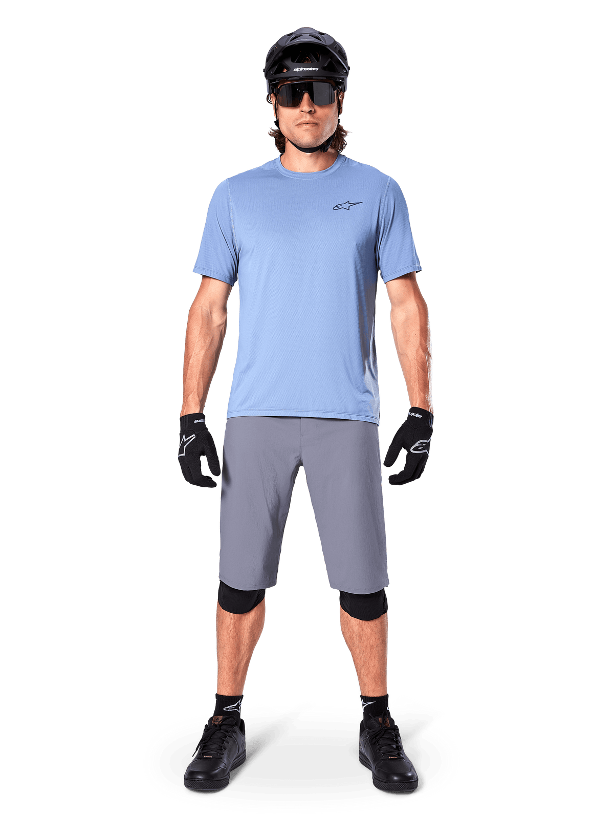 Mannelijke fietser die Alpinestars A-Dura Shorts in donkergrijs draagt, gecombineerd met een lichtblauw T-shirt met korte mouwen, zwarte helm, zonnebril en zwarte kniebeschermers, complete mountainbike-outfit voor off-road prestaties.