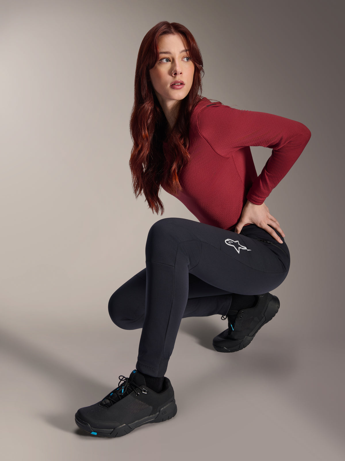 Alpinestars Stella A-Dura Thermal broek, MTB-broek voor dames, zwart, gedragen door een model in een gehurkte houding met een bordeauxrode top met lange mouwen en zwarte fietsschoenen, voorzien van een wit sterlogo op de dij, duurzame en warme constructie voor off-road ritten in koud weer