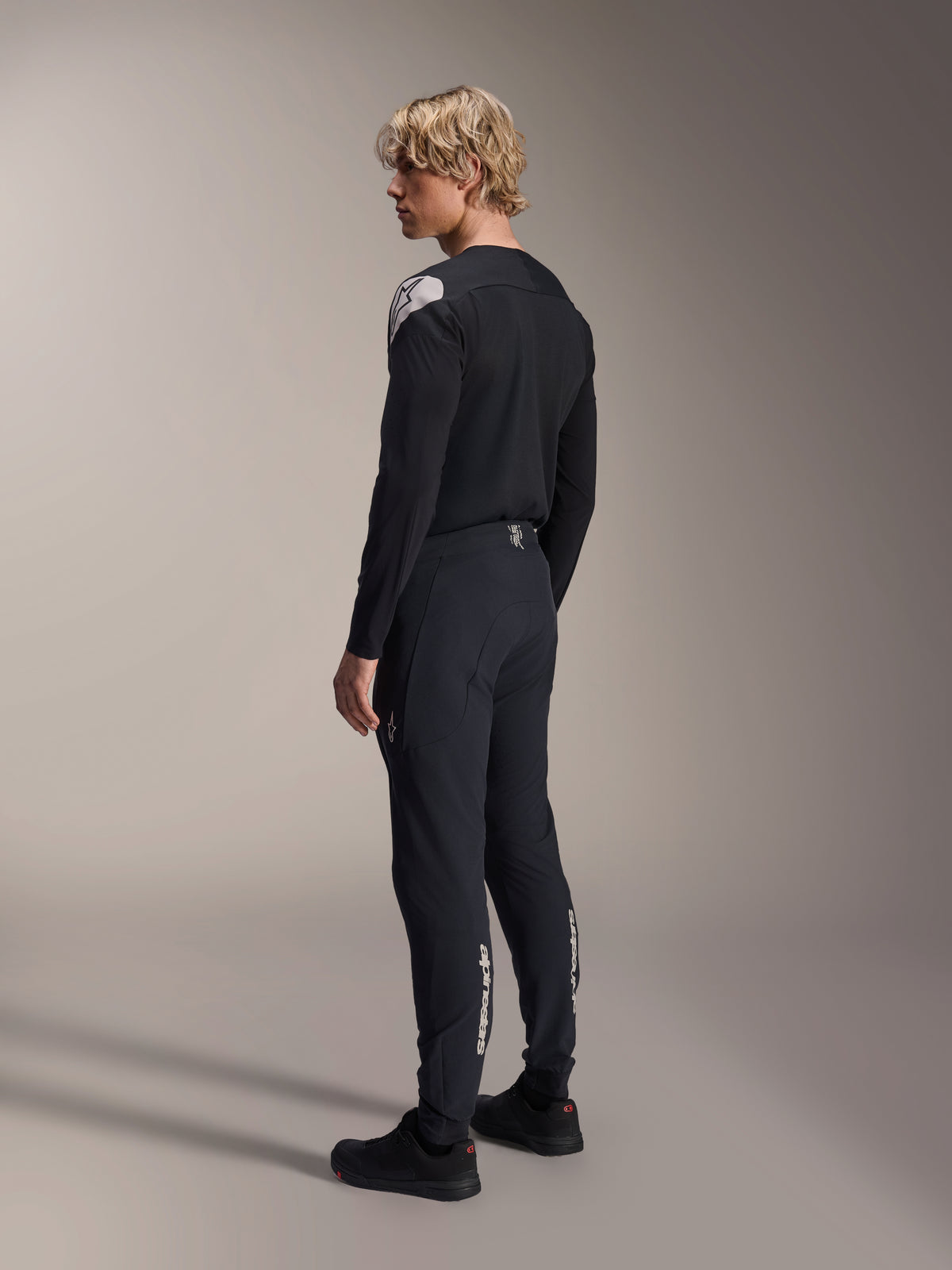 Alpinestars A-Aria Elite Hose, Mountainbike-Hose, Black Titanium, Dreiviertel-Rückansicht am Modell, mit weißem Alpinestars-Branding an den Unterschenkeln, Performance-Passform mit elastischen Bündchen, kombiniert mit einem passenden schwarzen Langarm-Trikot.