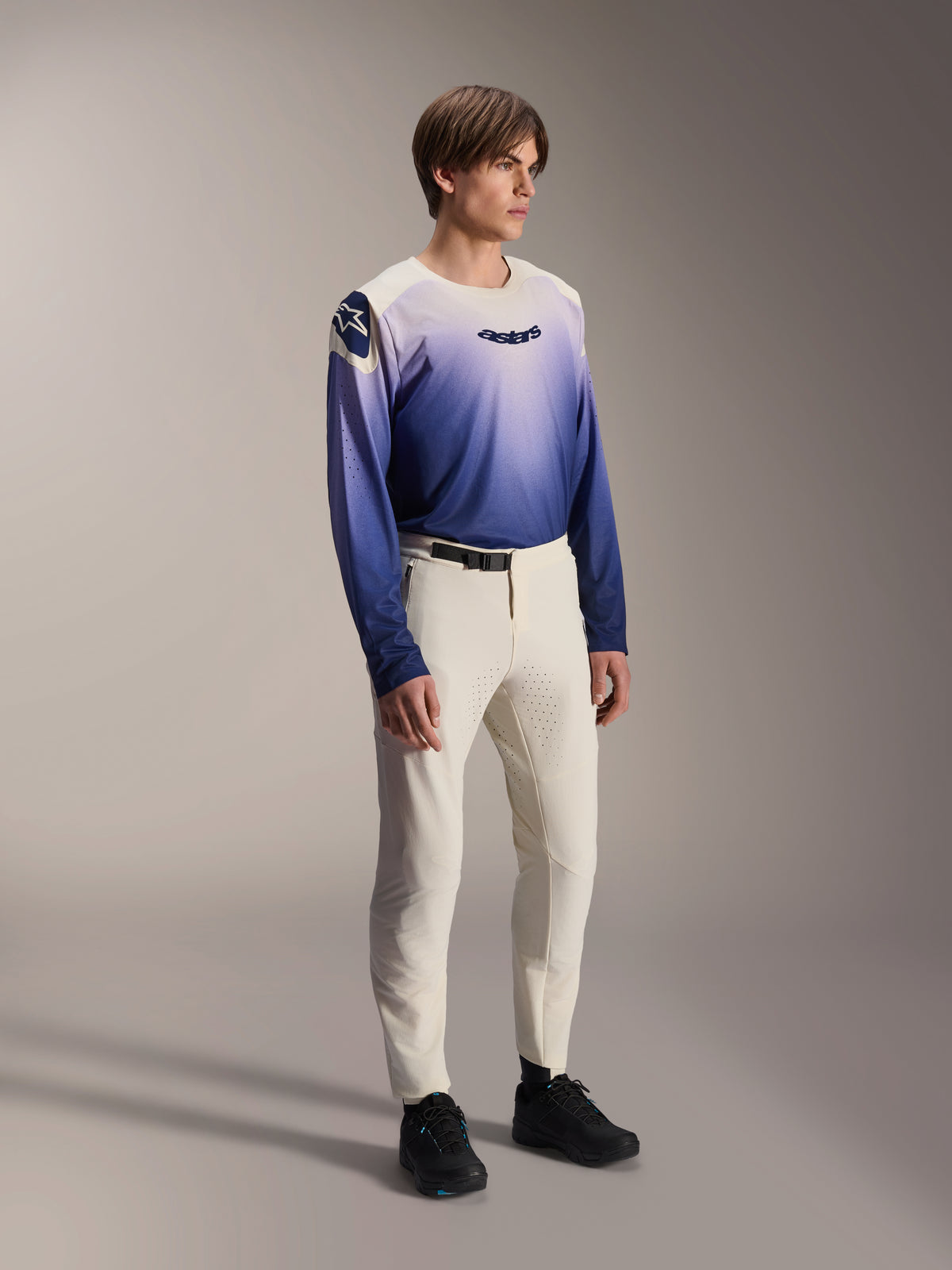 Model draagt Alpinestars A-Aria Elite Pants in Ecru, off-white mountainbikebroek met lasergesneden ventilatie en een zwarte gespsluiting, gecombineerd met een jersey met blauw en paars kleurverloop en lange mouwen, prestatiegerichte fietskleding voor off-road trails