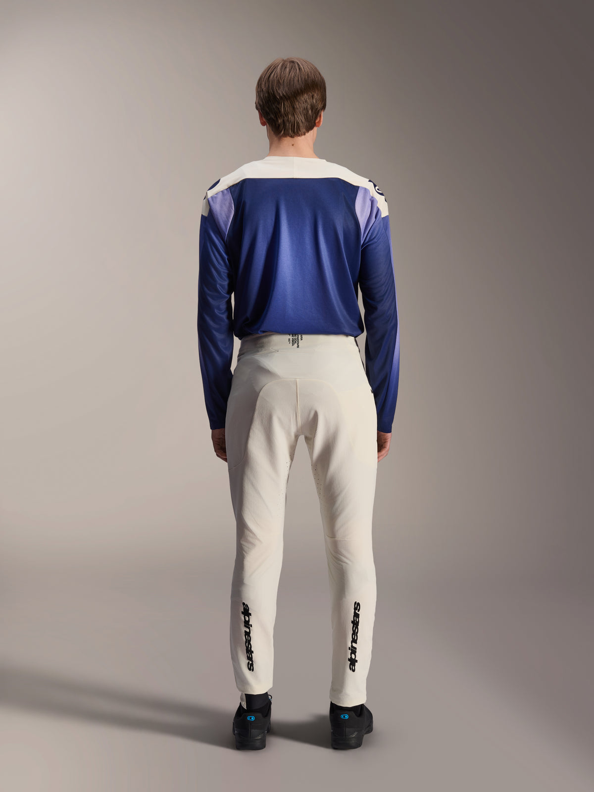 Alpinestars A-Aria Elite broek in Ecru, achteraanzicht op een model gecombineerd met een blauwe A-Aria Polartec Maker Elite jersey met lange mouwen, gebroken witte technische MTB-broek met zwarte logo's op de onderbenen, technische fietskleding voor mountainbiken
