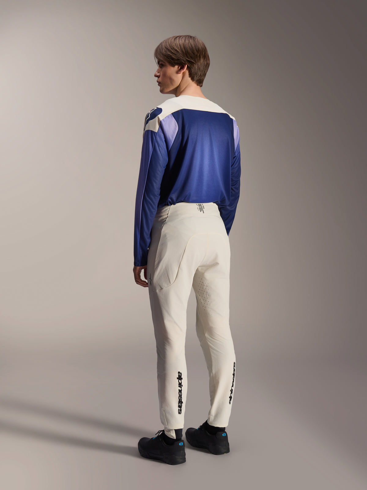 Alpinestars A-Aria Elite broek, MTB-broek, ecru gebroken wit, achteraanzicht gedragen door een model, voorzien van zwart woordmerk op de onderbenen en laserperforaties voor ventilatie, gecombineerd met een blauw en paars shirt met lange mouwen en zwarte schoenen, professionele mountainbike-uitrusting