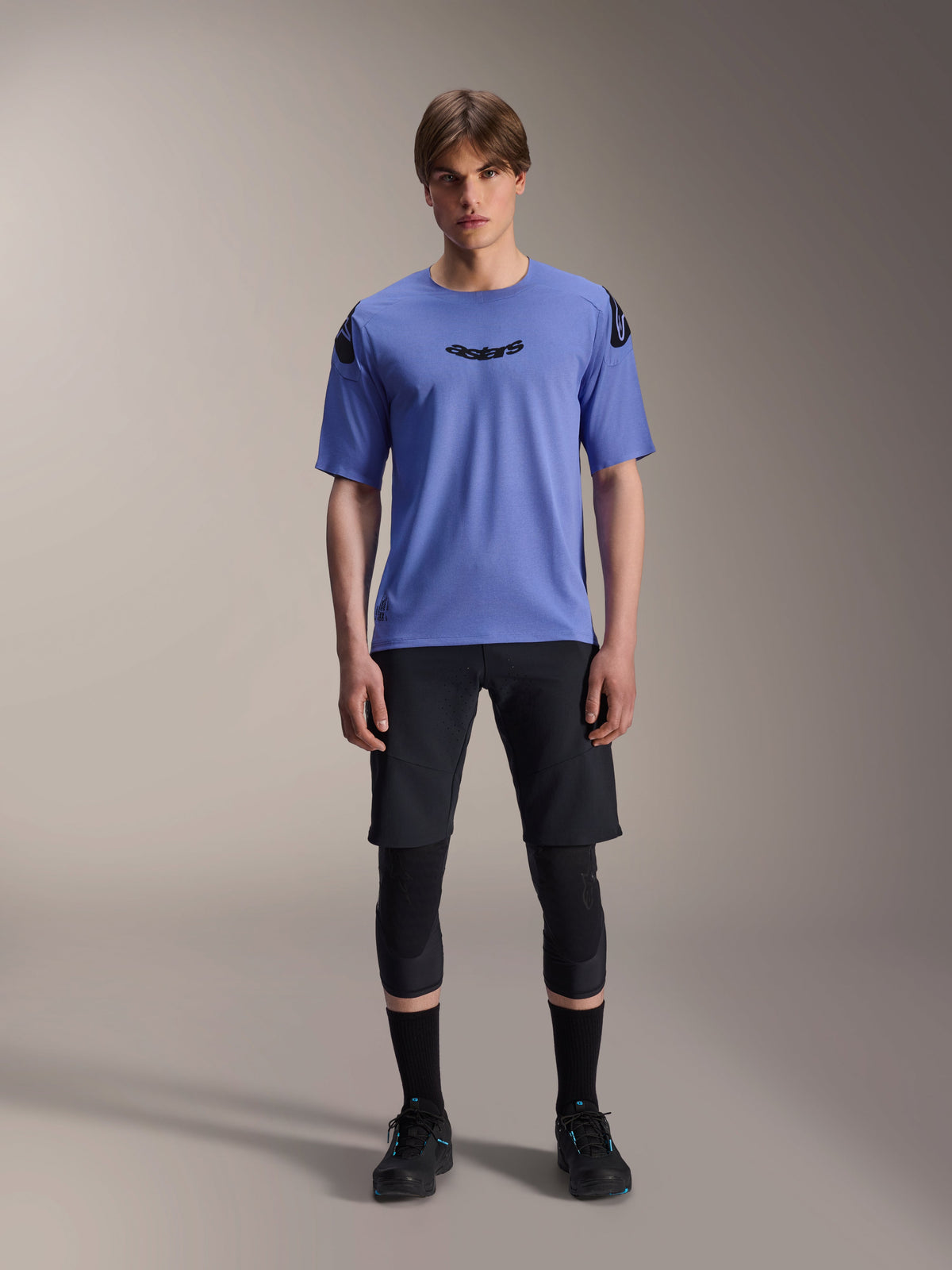 Mannelijk model draagt Alpinestars A-Aria Polartec Elite Jersey met korte mouwen in Baja Blue, zwart astars-logo op de borst, gecombineerd met zwarte MTB-short, kniebeschermers en zwarte fietsschoenen, studio-opname voor mountainbikekleding