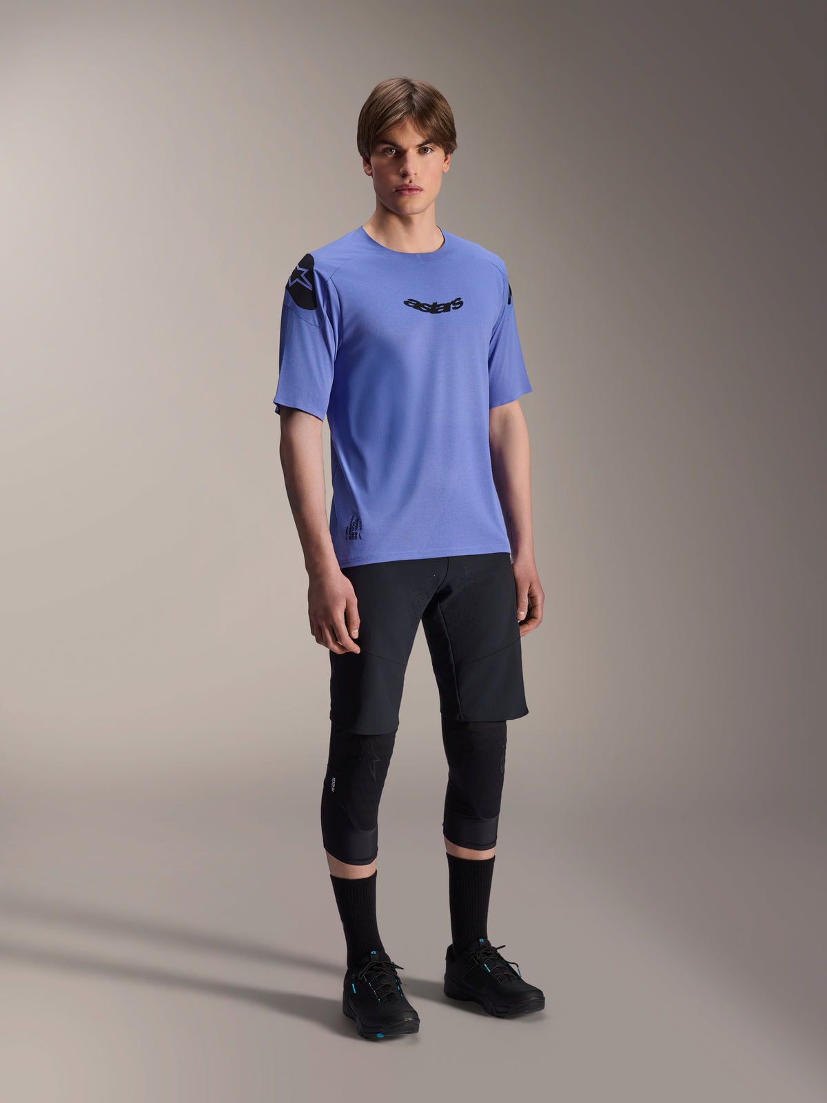 Mannelijk model draagt Alpinestars A-Aria Elite Shorts in Black Titanium en A-Aria Polartec Elite SS Jersey in lichtpaars, gecombineerd met zwarte mountainbike kniebeschermers en schoenen, professionele wielerkleding voor off-road prestaties.