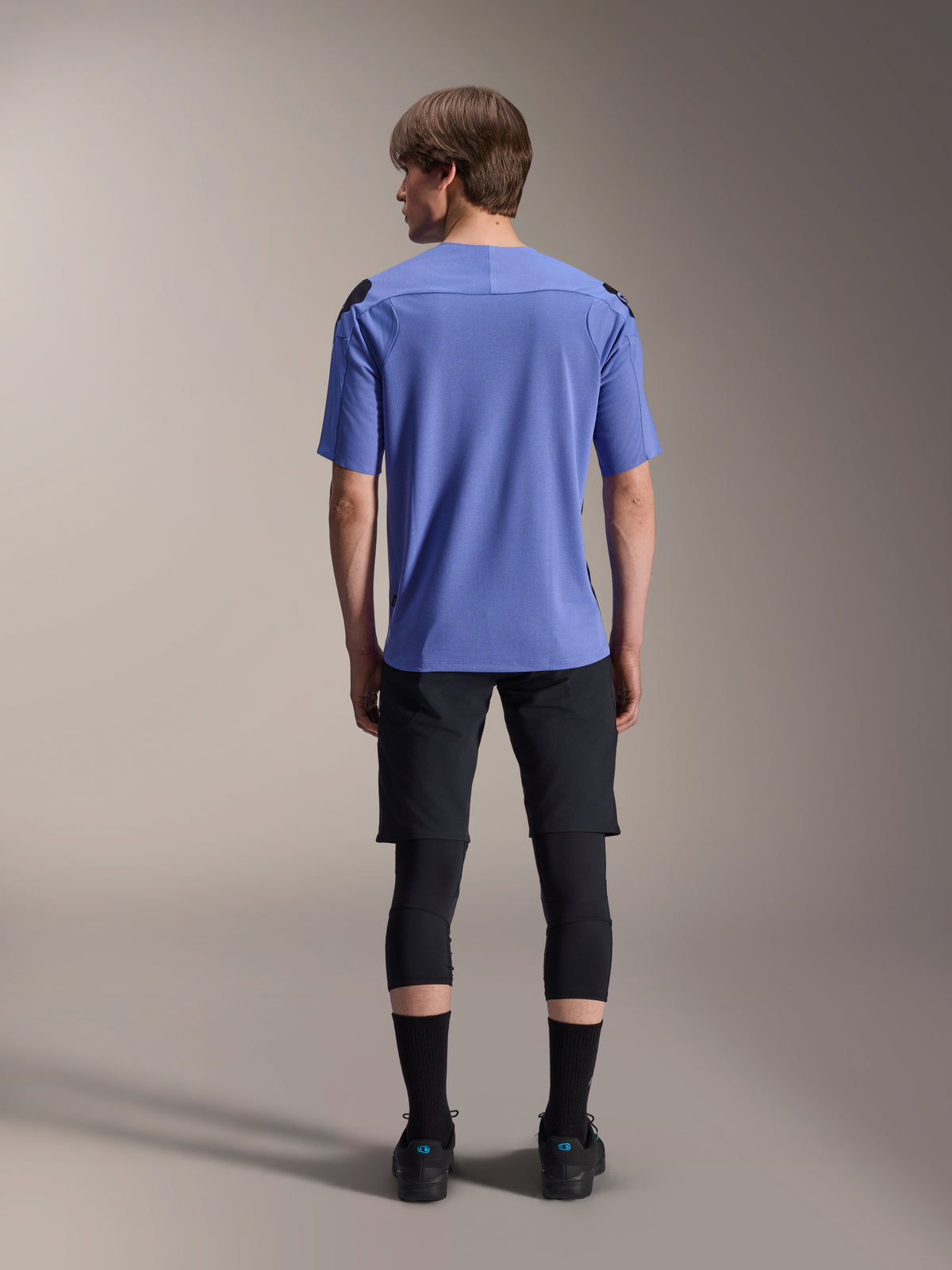 Mountainbike-Fahrer von hinten in Alpinestars A-Aria Lite Shorts in Schwarz, kombiniert mit einem blauen kurzärmeligen Trikot, schwarzen Knieschonern und schwarzen Socken, Performance-Radsportausrüstung für Offroad-Trails