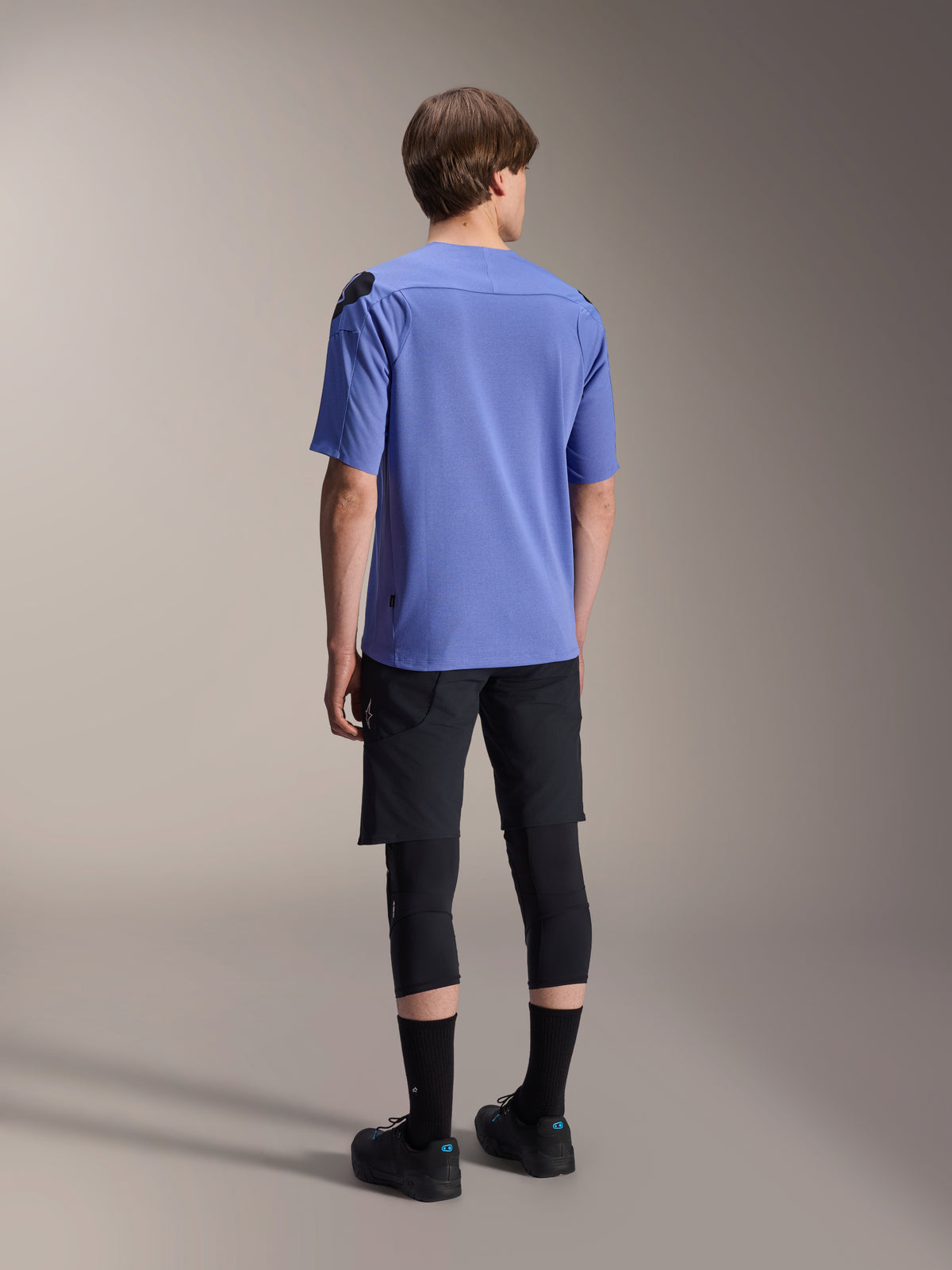 Alpinestars A-Aria Lite Shorts, schwarze Mountainbike-Shorts, kombiniert mit einem blauen A-Aria Polartec Elite Kurzarm-Trikot und schwarzen Knieschonern, Rückansicht eines Fahrers in leistungsstarker Fahrradausrüstung für Offroad-Trails.
