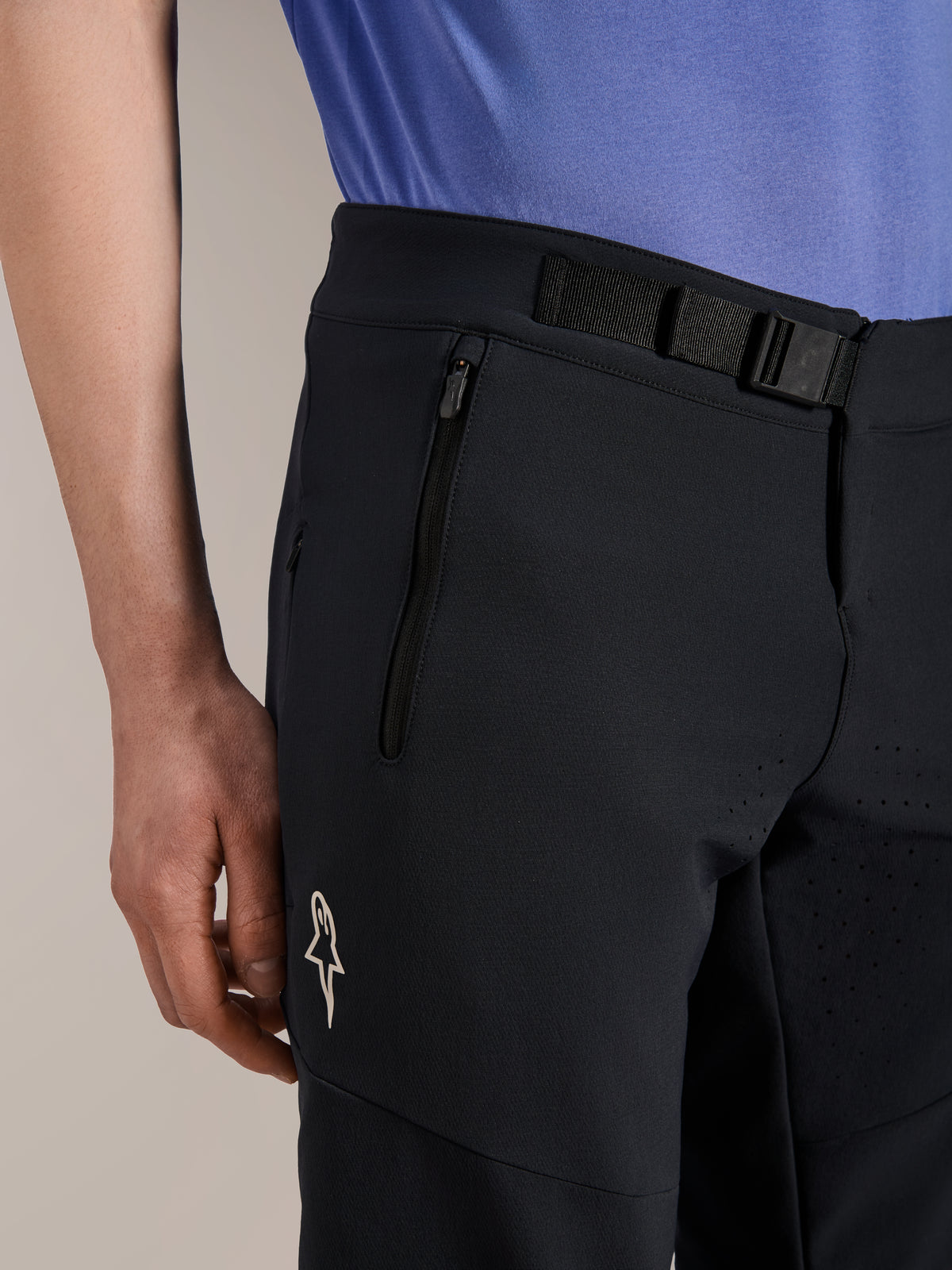 Alpinestars A-Aria Elite Shorts, mountainbikebroek, Black Titanium, voorzien van een magnetische gespsluiting, zak met rits en wit Alpinestars-logo, gedragen met een blauw fietsshirt, hoogwaardige mountainbike-uitrusting