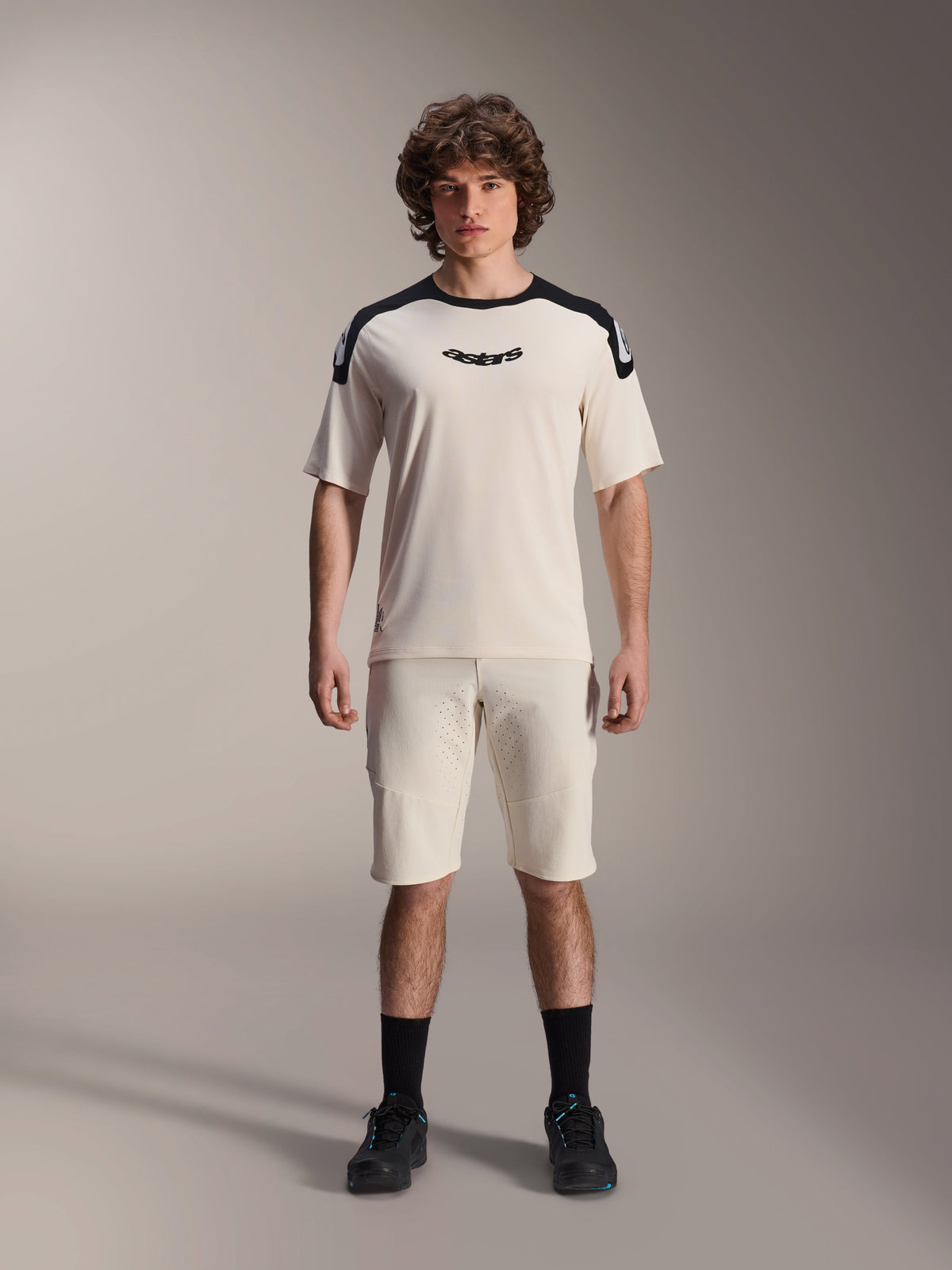 Alpinestars A-Aria Polartec Elite Jersey met korte mouwen in Ecru, voorzien van zwarte schouderpanelen en een centraal borstlogo, gecombineerd met bijpassende ecru short, zwarte sokken en fietsschoenen, hoogwaardige mountainbike-uitrusting.