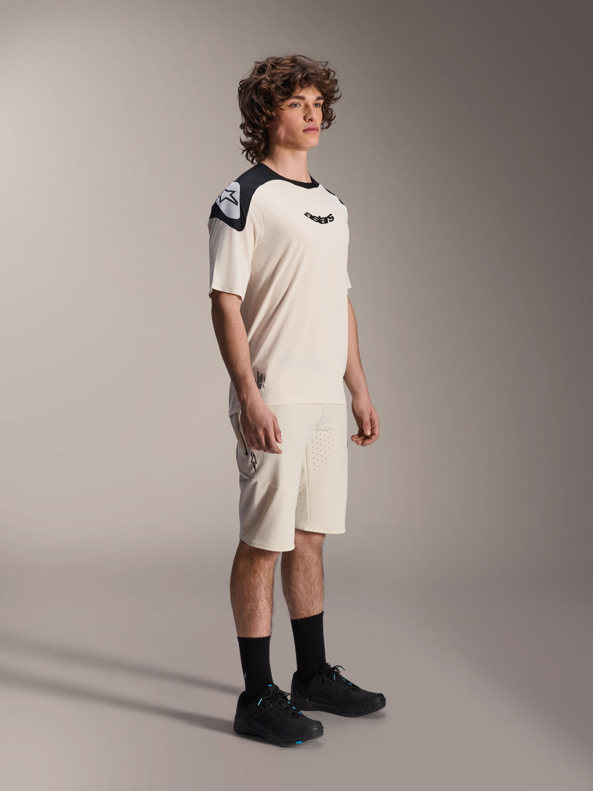 Alpinestars A-Aria Polartec Elite Jersey met korte mouwen, ecru met zwarte schouderpanelen en logo, gedragen door een model met bijpassende shorts en zwarte schoenen, hoogwaardige mountainbikekleding voor off-road trailrijden.