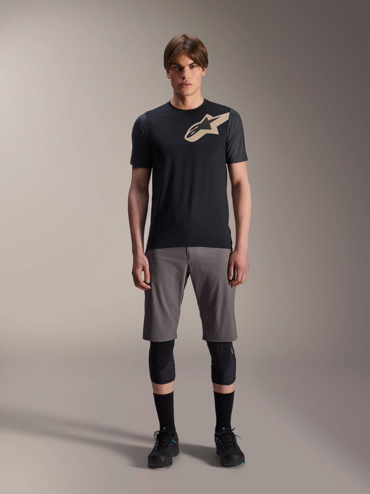 Maillot à manches courtes Alpinestars A-Dura Lipan, noir avec manches gris chiné et logo Astars beige sur la poitrine, porté par un mannequin homme avec un short de VTT gris et des genouillères noires, équipement de performance pour le VTT