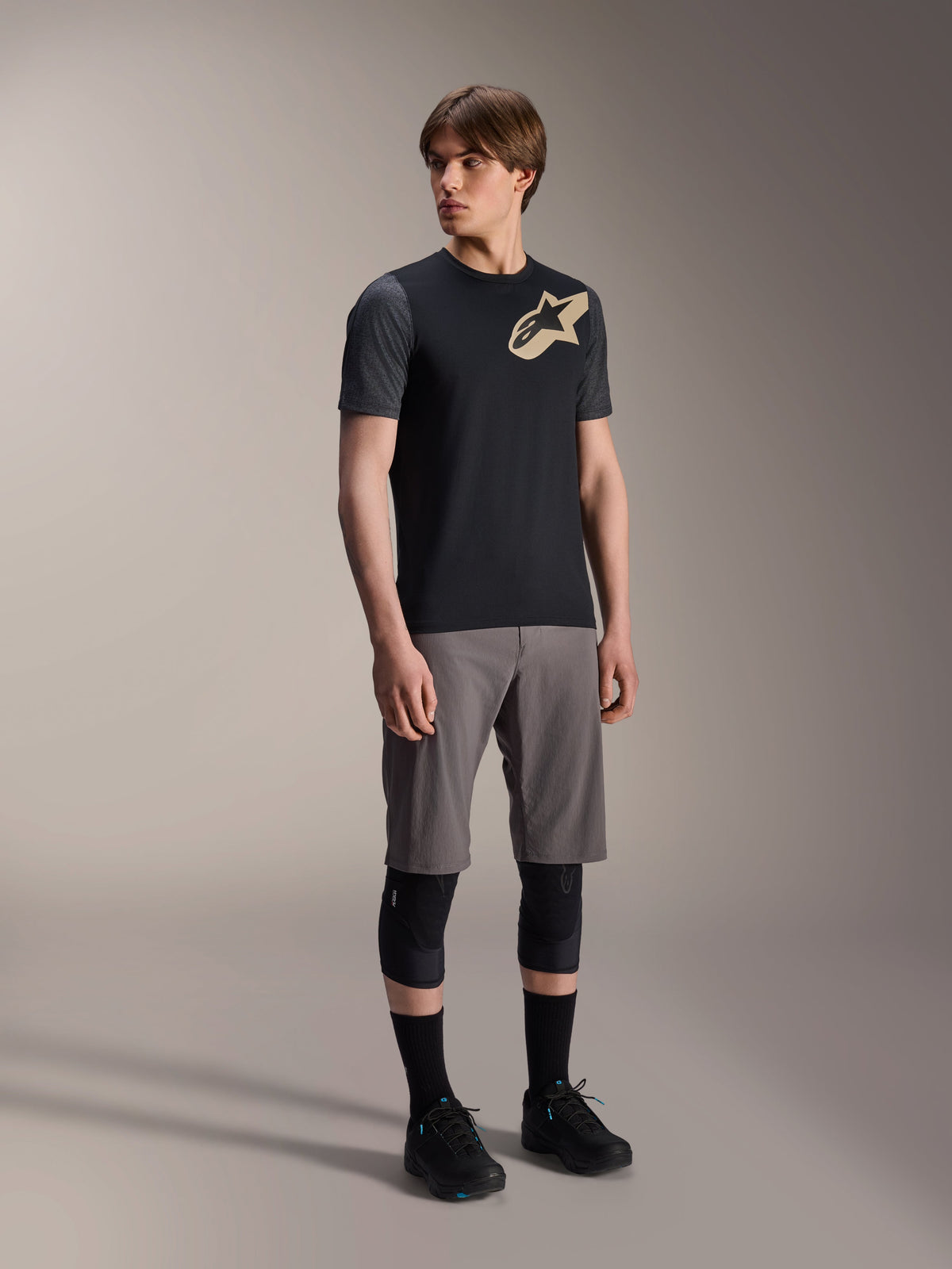 Mannequin homme portant le maillot à manches courtes Alpinestars A-Dura Lipan en noir, avec des manches gris foncé et un logo Astars beige sur la poitrine, associé à un short de VTT gris, des genouillères noires et des chaussures de cyclisme noires, tenue de VTT coordonnée
