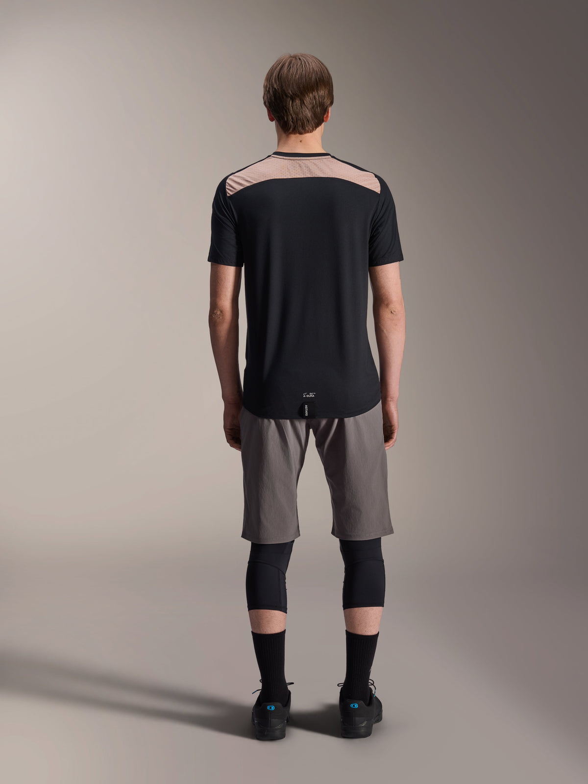 Maillot à manches courtes Alpinestars A-Dura Lipan, noir avec empiècement beige sur le haut du dos, vue arrière montrant l'ourlet arrière allongé, porté avec un short de VTT gris et des genouillères noires, vêtement de performance pour le VTT