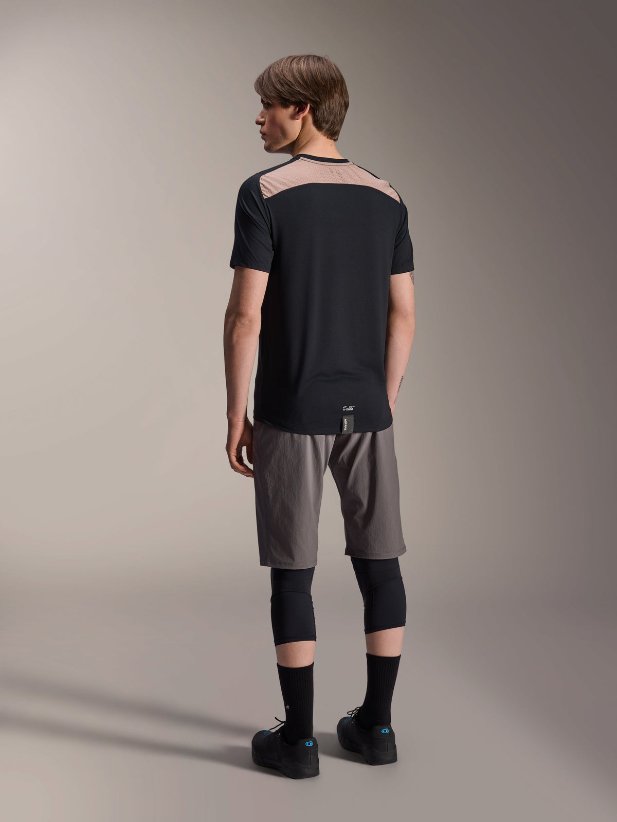 Maillot à manches courtes Alpinestars A-Dura Lipan noir, vue de dos sur mannequin présentant l'empiècement en mesh perforé marron clair sur le haut du dos, associé à un short de VTT gris et des genouillères noires pour une tenue de trail complète.