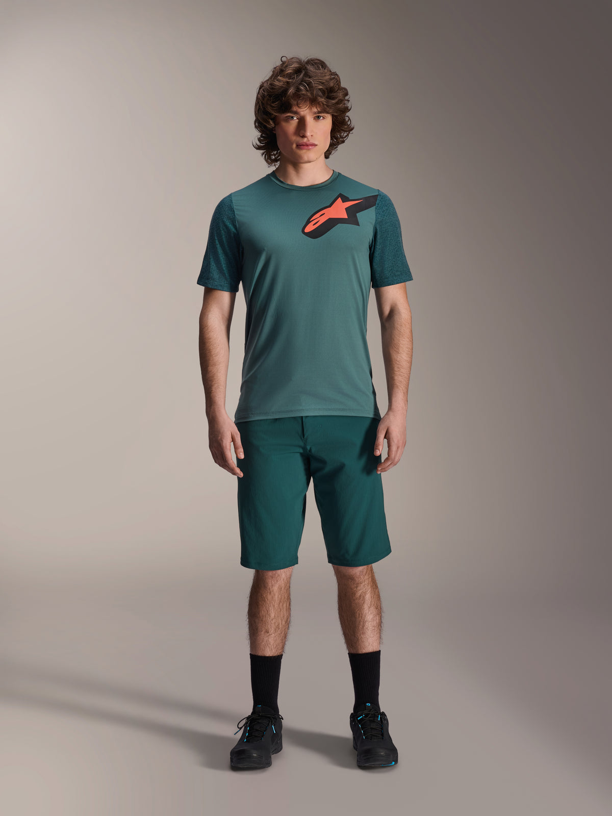 Alpinestars A-Dura Shorts in Dark Pine, Herren-Mountainbike-Shorts, an einem Modell mit passendem Kurzarmtrikot in Teal mit rot-schwarzem Stern-Logo, schwarze Socken und Radschuhe, lässige sportliche Mountainbike-Bekleidung