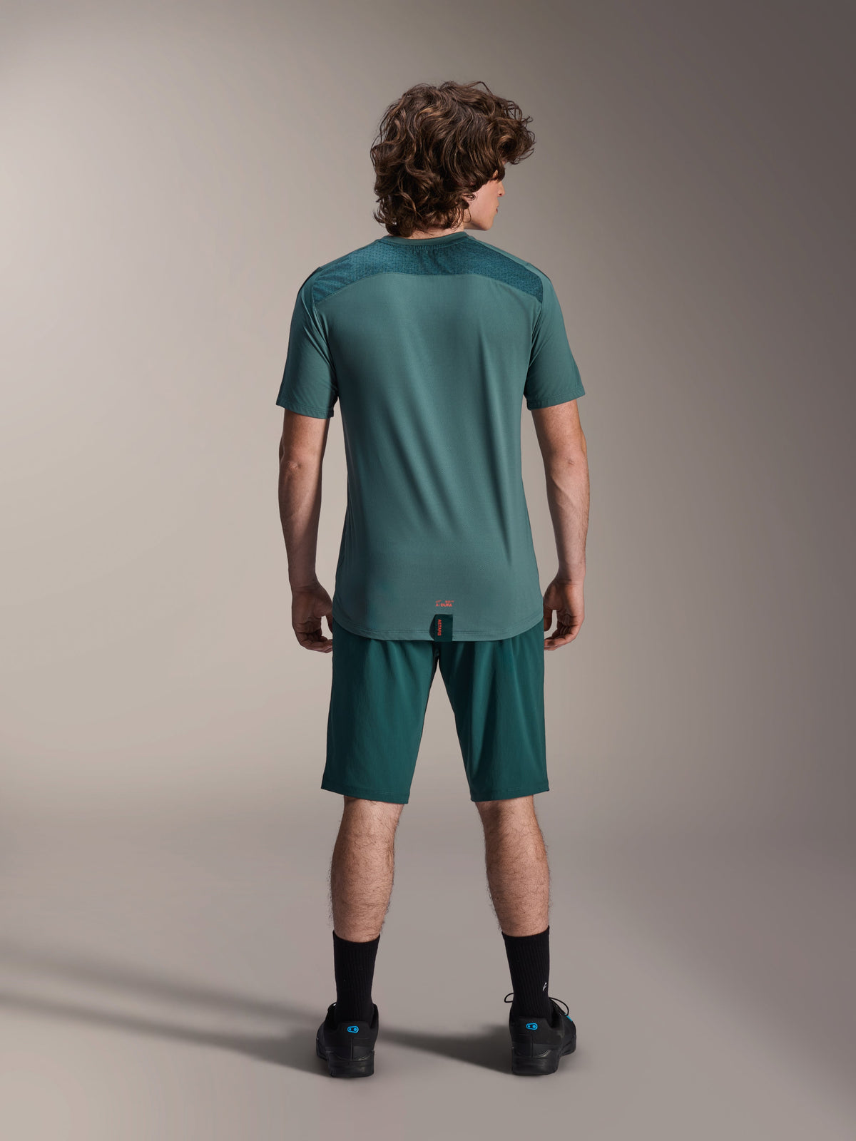 Maillot à manches courtes Alpinestars A-Dura Lipan en Silver Pine, vue de dos montrant les empiècements en jacquard texturé aux épaules et l'ourlet arrière plus long, porté avec un short de VTT vert foncé et des chaussures noires.
