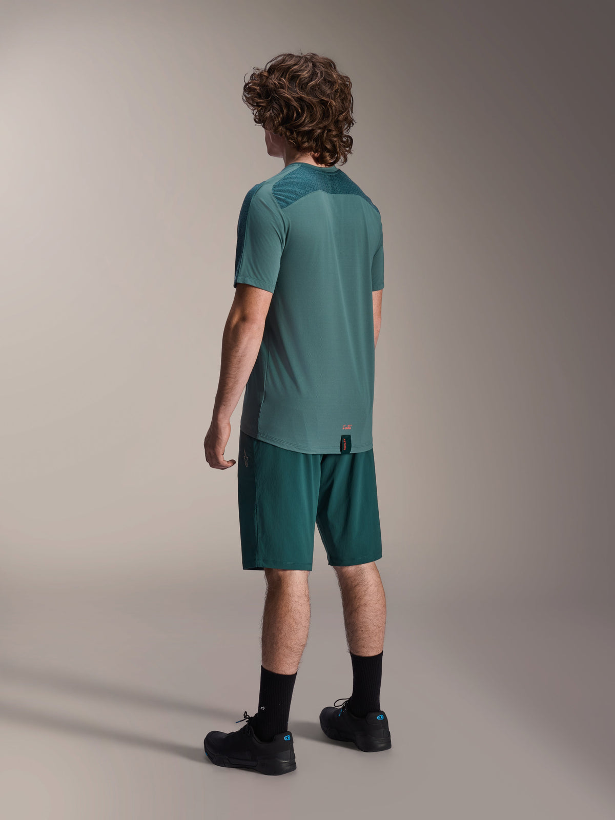 Modèle masculin portant le maillot à manches courtes Alpinestars A-Dura Lipan en Silver Pine, vu de dos, associé à un short de VTT vert foncé assorti, des chaussettes et chaussures noires, avec un empiècement texturé sur le haut du dos et un ourlet arrière allongé pour une meilleure couverture.
