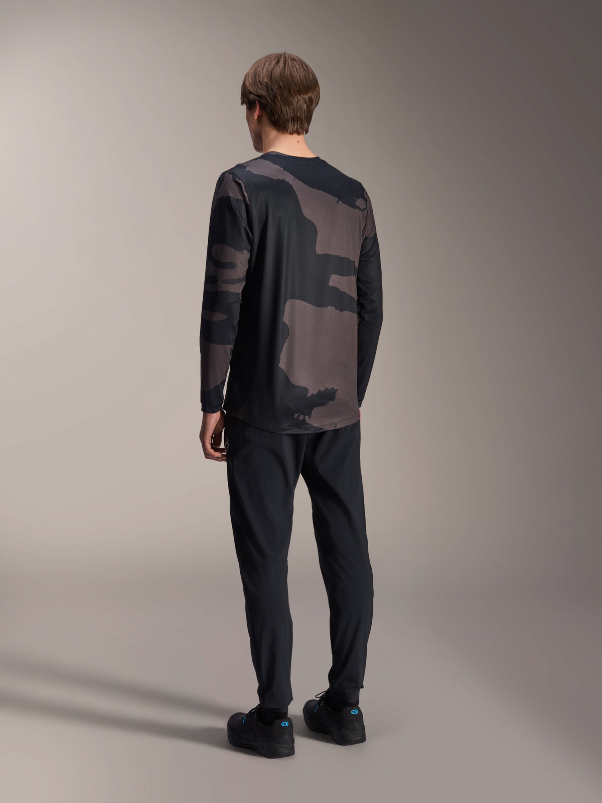 Alpinestars A-Dura Hollow Camo Langarm-Trikot, Black Camo, Rückansicht des Modells, technisches Mountainbike-Trikot mit verlängertem Rückensaum, kombiniert mit schwarzer Hose und Radschuhen, lässige Offroad-Performance-Ausrüstung