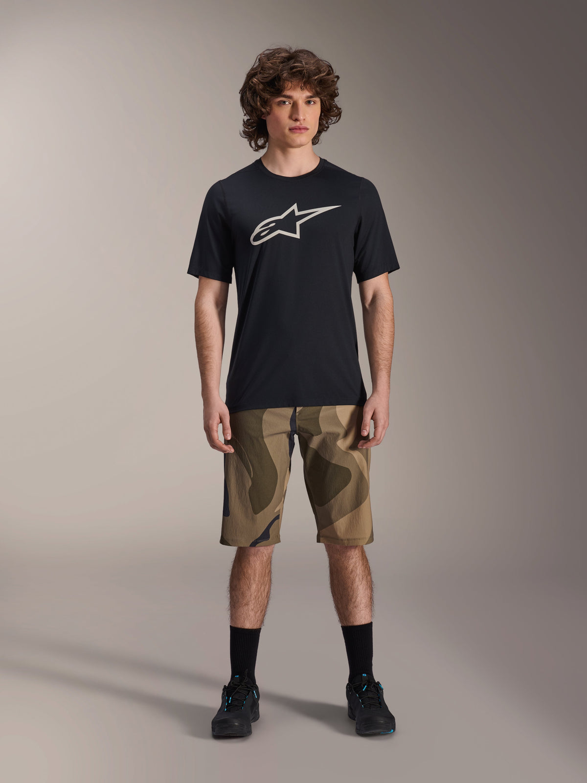 Alpinestars A-Dura Astar jersey met korte mouwen, zwart met groot wit Astars-logo op de borst, gedragen door een model met camouflage MTB-short en zwarte fietsschoenen, casual mountainbike-kleding
