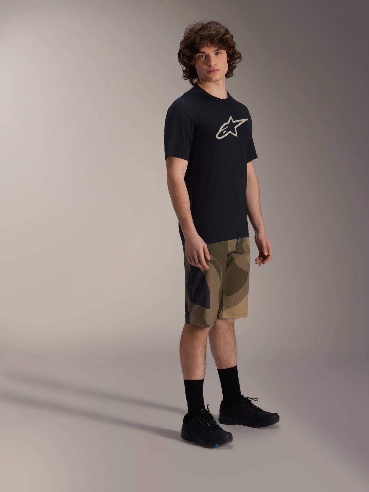 Alpinestars A-Dura Astar Jersey met korte mouwen, zwart met wit logo op de borst, gedragen met camouflage short en zwarte MTB-schoenen, casual mountainbike-uitrusting