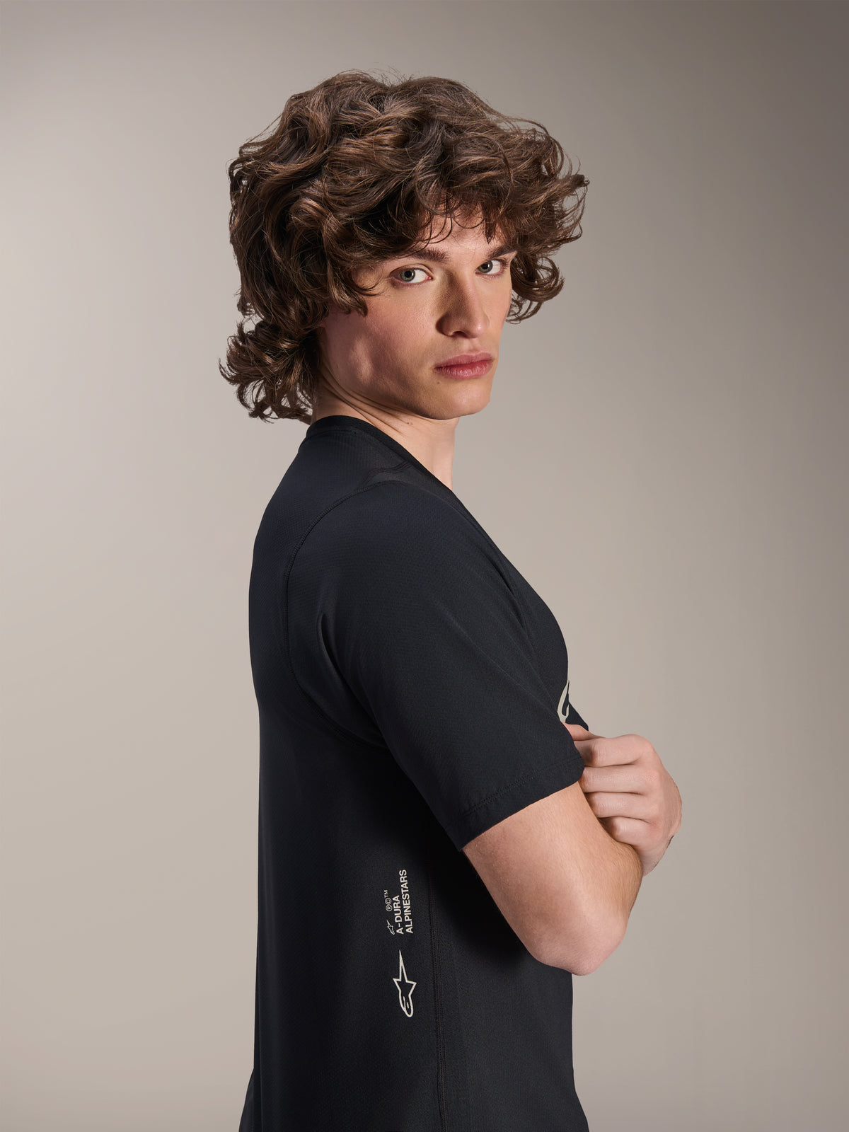 Alpinestars A-Dura Astar Jersey, MTB-shirt met korte mouwen, zwart, mannelijk model poserend in profiel met de armen over elkaar, voorzien van witte Alpinestars-branding aan de onderzijde, casual sportief ontwerp voor mountainbiken.