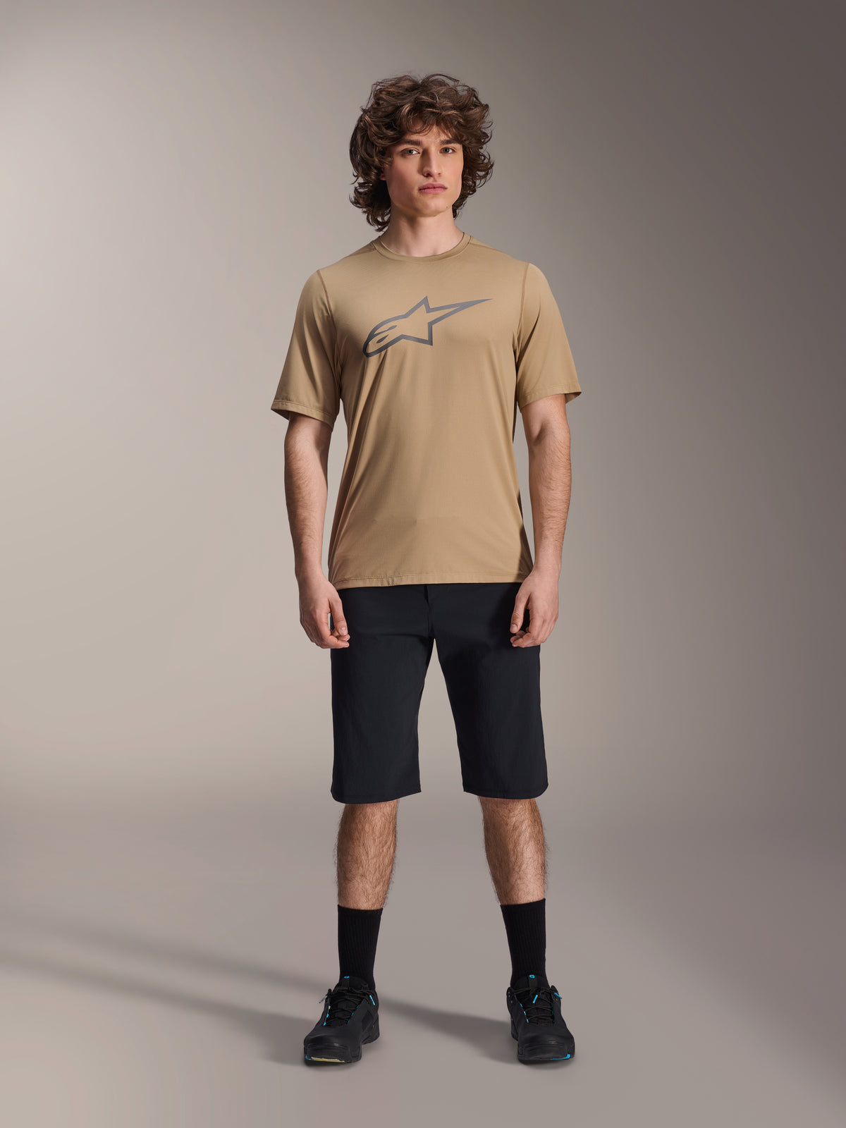 Alpinestars A-Dura Liner Shorts, MTB-shorts, zwart, gedragen met een lichtbruine jersey met korte mouwen en grijs ster-logo, zwarte sokken en mountainbikeschoenen, casual sportieve stijl