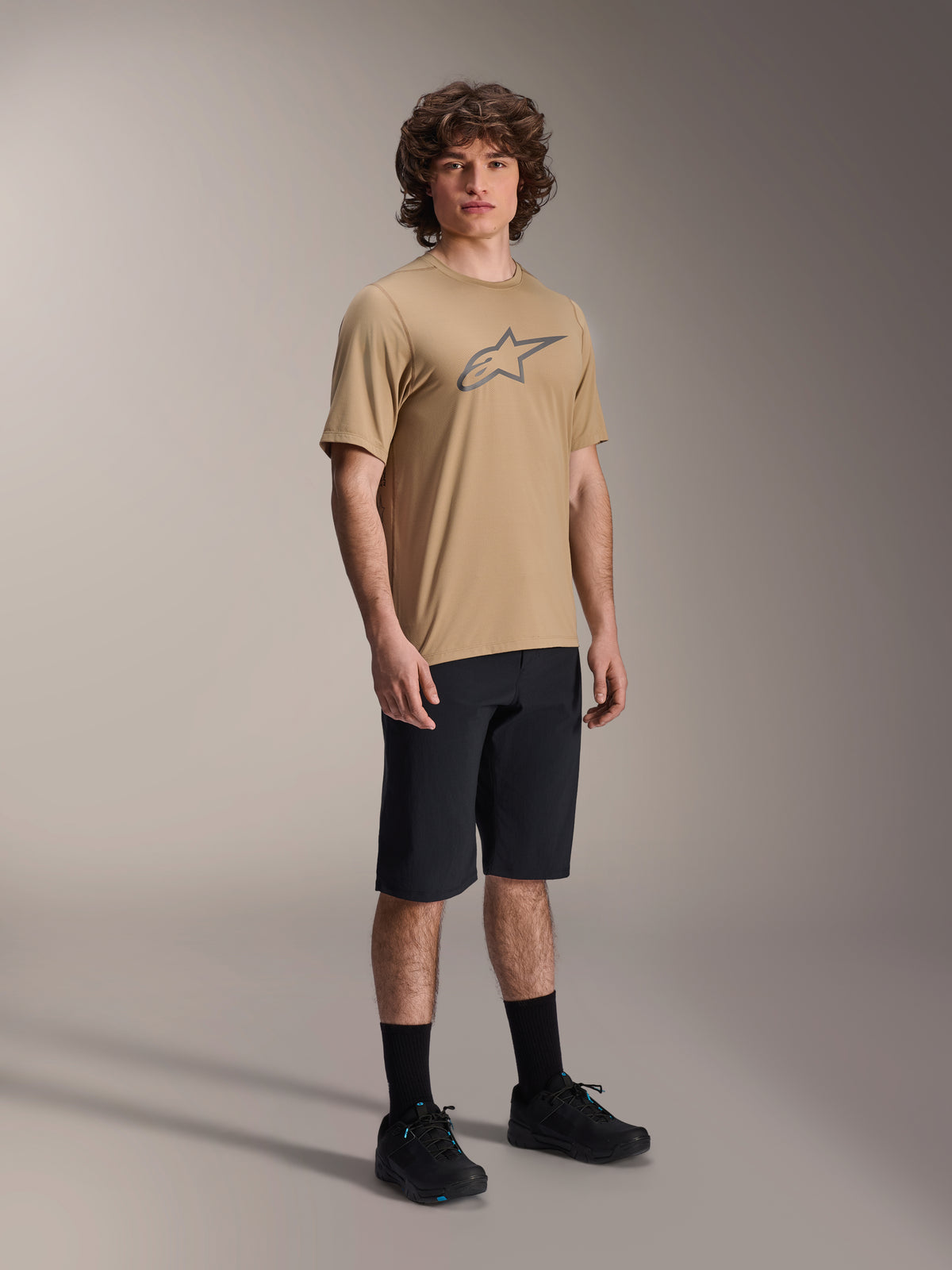 Mannelijk model in zwarte Alpinestars A-Dura Liner Shorts, gecombineerd met een beige A-Dura SS Jersey met grijs Astars-logo, zwarte fietssokken en mountainbikeschoenen, complete off-road fietsoutfit