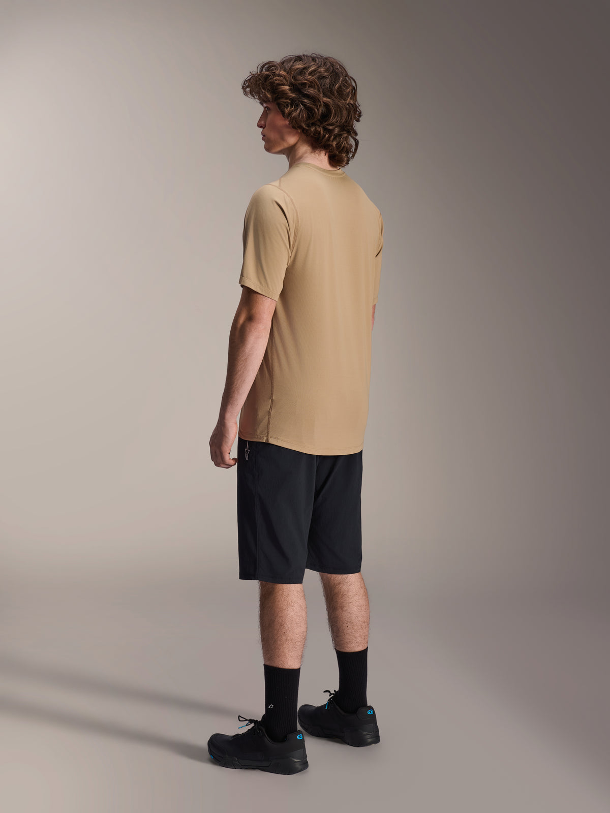 Alpinestars A-Dura Liner Shorts in zwart en A-Dura SS Jersey in tan, gedragen door een mannelijk model in zijaanzicht, gecombineerd met zwarte sokken en fietsschoenen, mountainbike lifestyle kleding.