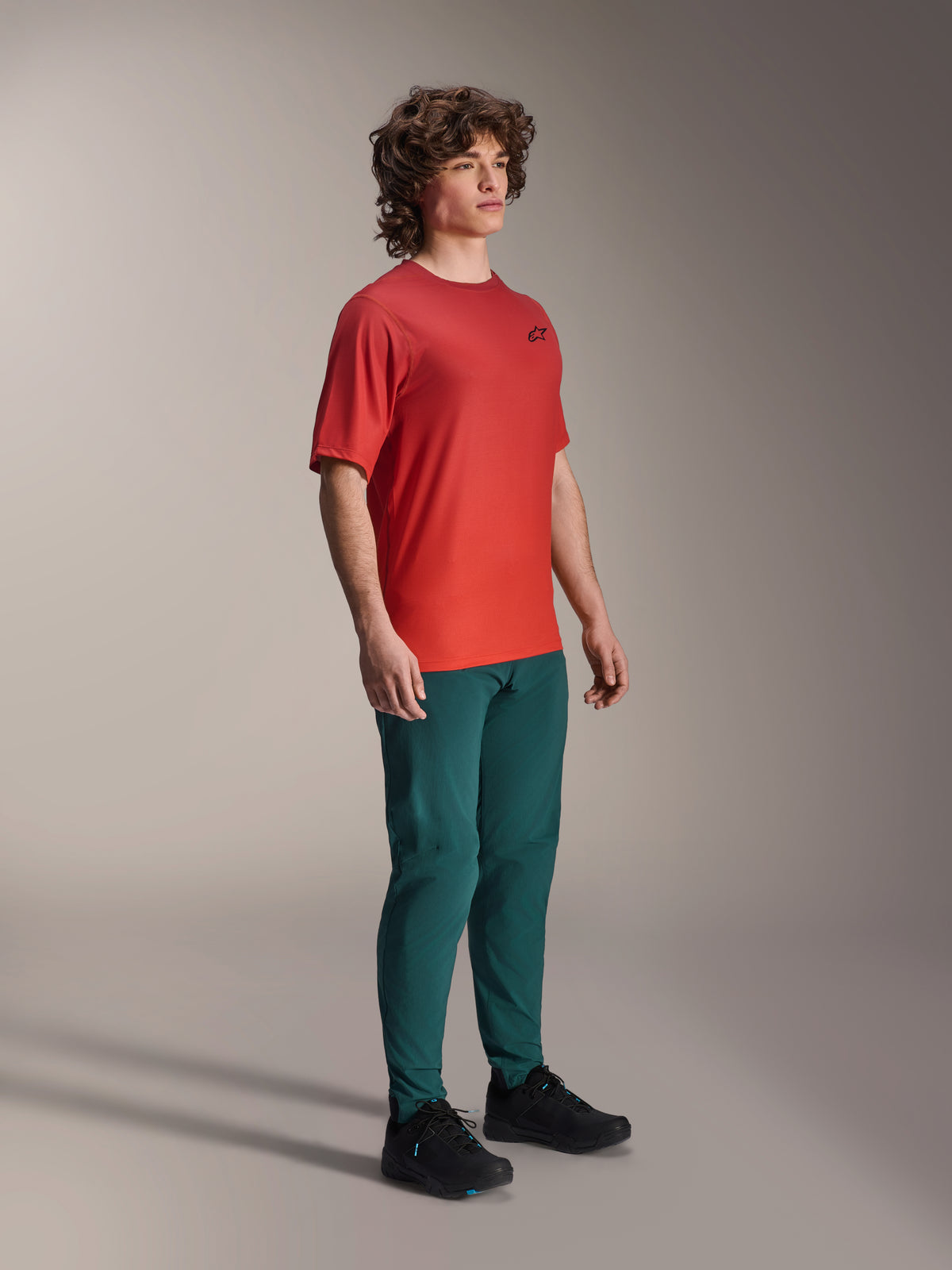 Model draagt Alpinestars A-Dura Pants in Dark Pine, gecombineerd met een rood A-Dura Rise Jersey met korte mouwen en zwarte MTB-schoenen, sportieve mountainbike-kleding in een studio-omgeving