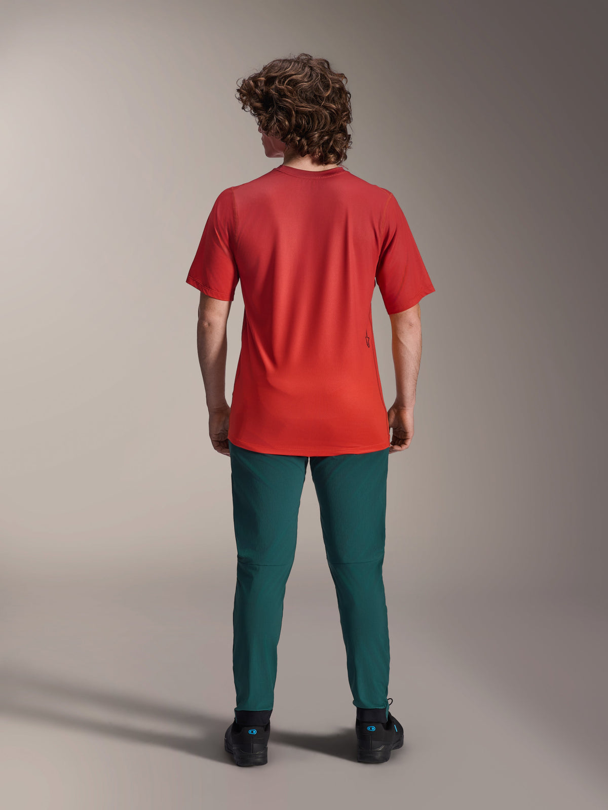 Alpinestars A-Dura Rise Kurzarmtrikot, Spicy Orange, Rückansicht an einem Model mit dunkeltürkiser Hose und schwarzen Schuhen, mit verlängertem Rückensaum und einem kleinen Siebdruck-Logo auf der Rückseite, leichte Performance-MTB-Ausrüstung