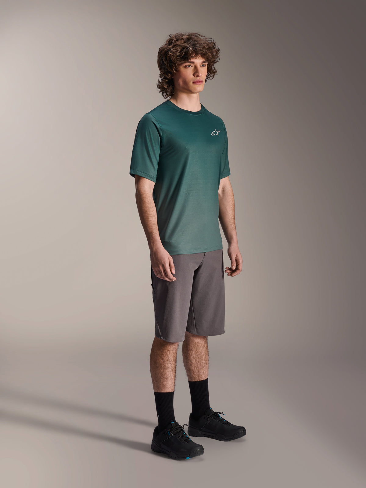Alpinestars A-Dura Rise Jersey - Short Sleeve en Silver Pine, maillot de VTT vert porté par un mannequin avec un short gris et des baskets noires, tenue de VTT décontractée avec un petit logo blanc sur la poitrine