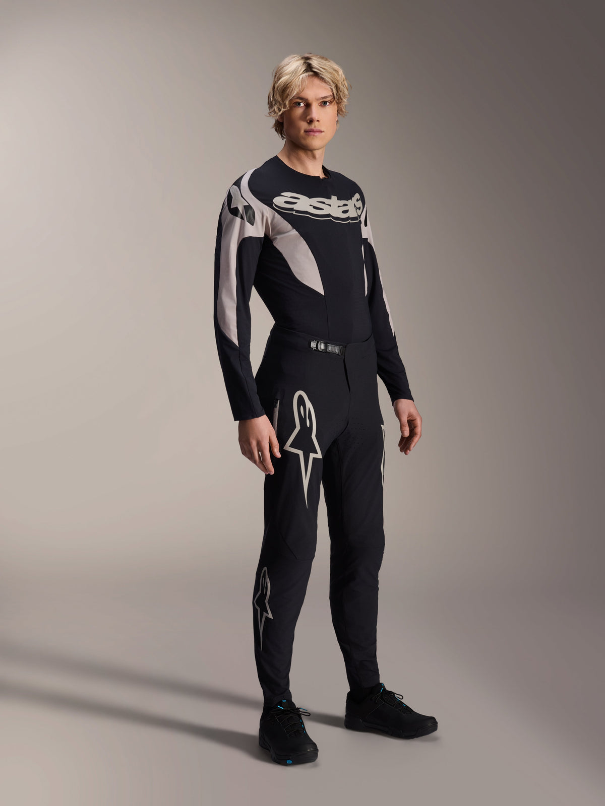 Modèle masculin portant le maillot Alpinestars A-Supra Melt à manches longues, noir avec des accents graphiques gris, logo 'astars' sur la poitrine, associé à un pantalon et des chaussures noirs assortis, équipement de VTT haute performance pour la course tout-terrain