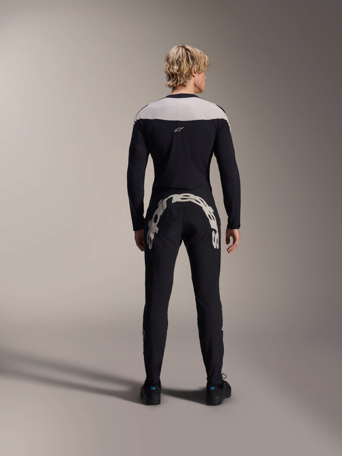 Homme vu de dos portant le maillot à manches longues Alpinestars A-Supra Melt noir avec empiècement gris clair aux épaules, associé à un pantalon de VTT noir arborant un large logo Alpinestars à l'arrière, tenue de VTT haute performance