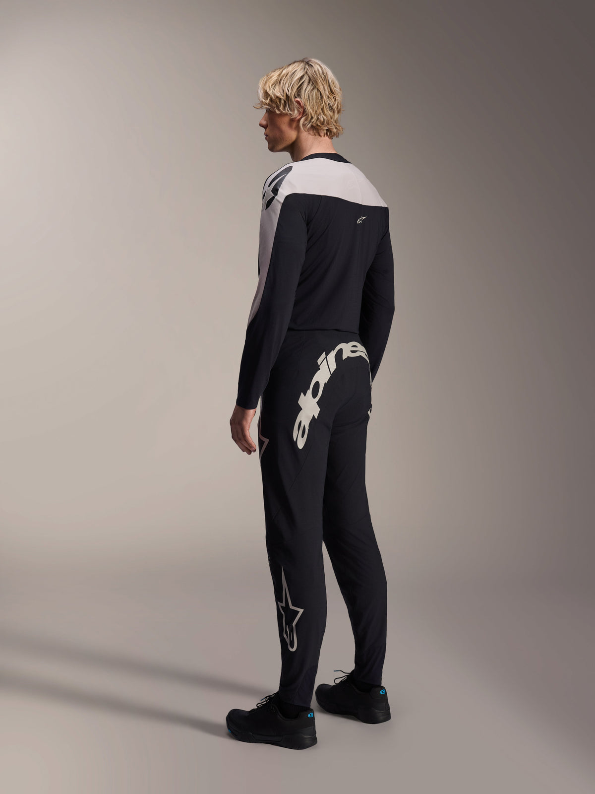Maillot à manches longues Alpinestars A-Supra Melt, noir avec empiècements blancs aux épaules, vu de dos et de côté sur un modèle, associé à un pantalon de VTT noir avec un grand logo blanc, tenue de course VTT haute performance