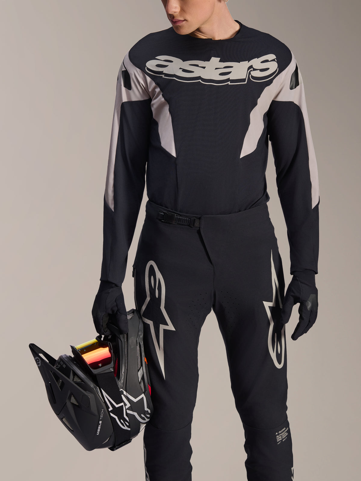Maillot à manches longues Alpinestars A-Supra Melt, noir avec empiècements graphiques gris et marquage 'astars' sur la poitrine, porté avec un pantalon de VTT et des gants noirs tout en tenant un casque intégral noir, tenue de course VTT haute performance