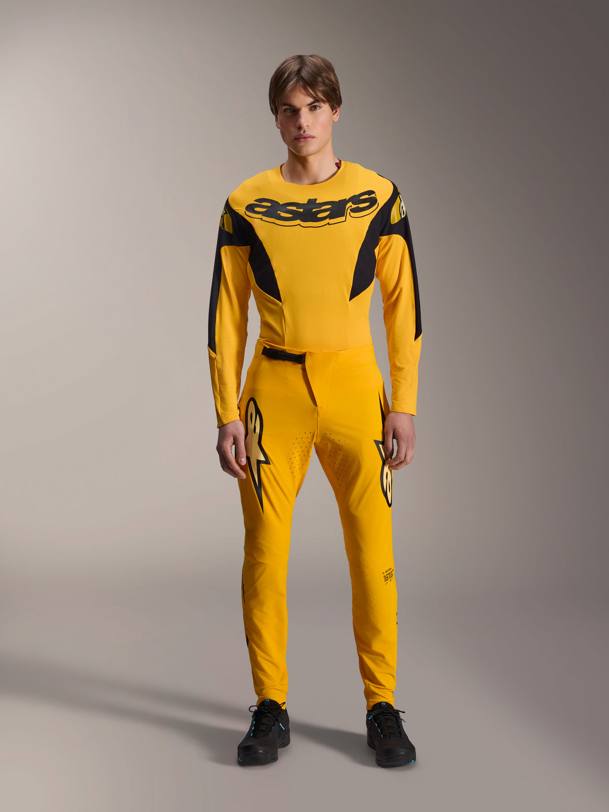 Limited Edition A-Supra Melt Gold-uitrustingsset, Alpinestars mountainbikekleding, donkergeel en zwart, model draagt jersey met lange mouwen met opvallend 'astars'-logo op de borst en bijpassende performance-broek, voorzien van lasergesneden ventilatie op de binnendijen en een veilige ratelsluiting, professionele MTB-racekit
