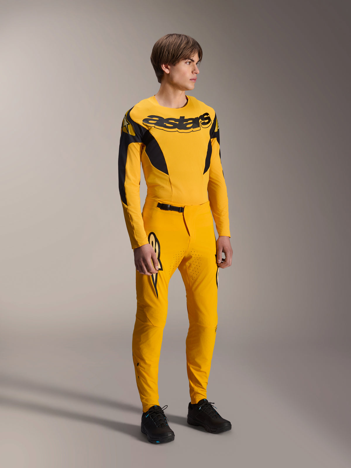 Mannelijk model draagt de Alpinestars Limited Edition A-Supra Melt Gold gear-set, gele MTB-jersey met lange mouwen en opvallende zwarte branding op de borst, bijpassende gele broek met lasergesneden ventilatie en zwarte accenten, complete mountainbike race-uitrusting