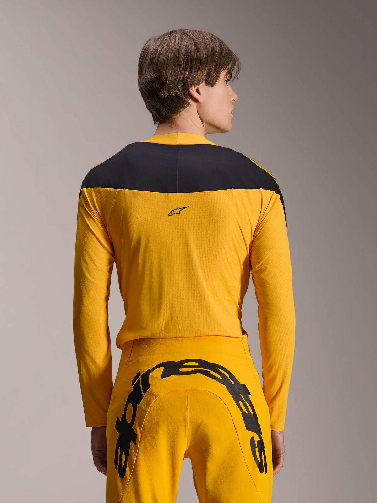 Alpinestars Limited Edition A-Supra Melt Gold Gear-Set, gele MTB-kledingset, achteraanzicht van model met jersey met lange mouwen en zwart schouderpaneel en bijpassende gele broek met groot zwart logo op het achterpand, hoogwaardige fietskleding.