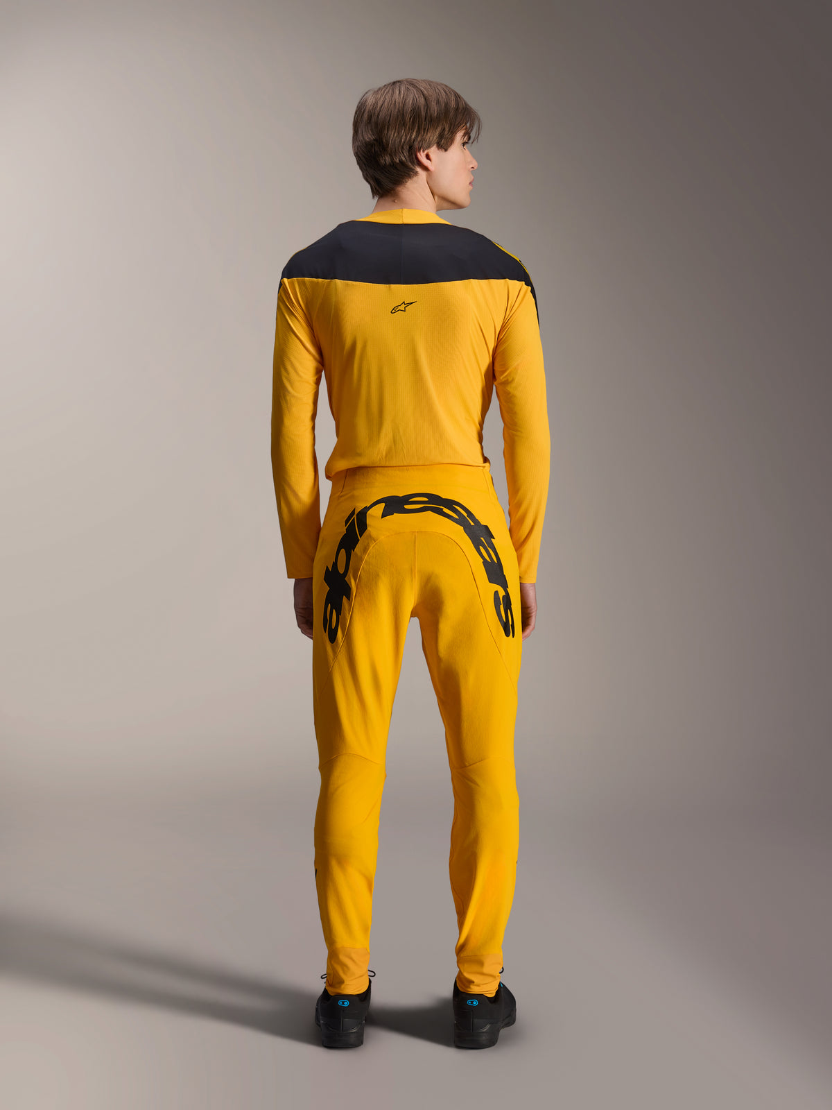 Alpinestars Limited Edition A-Supra Melt Gold Gear-Set, achteraanzicht van een model in donkergeel MTB-shirt met lange mouwen en zwart schouderstuk, donkergele broek met groot zwart Alpinestars-woordmerk op het achterpand, zwarte schoenen