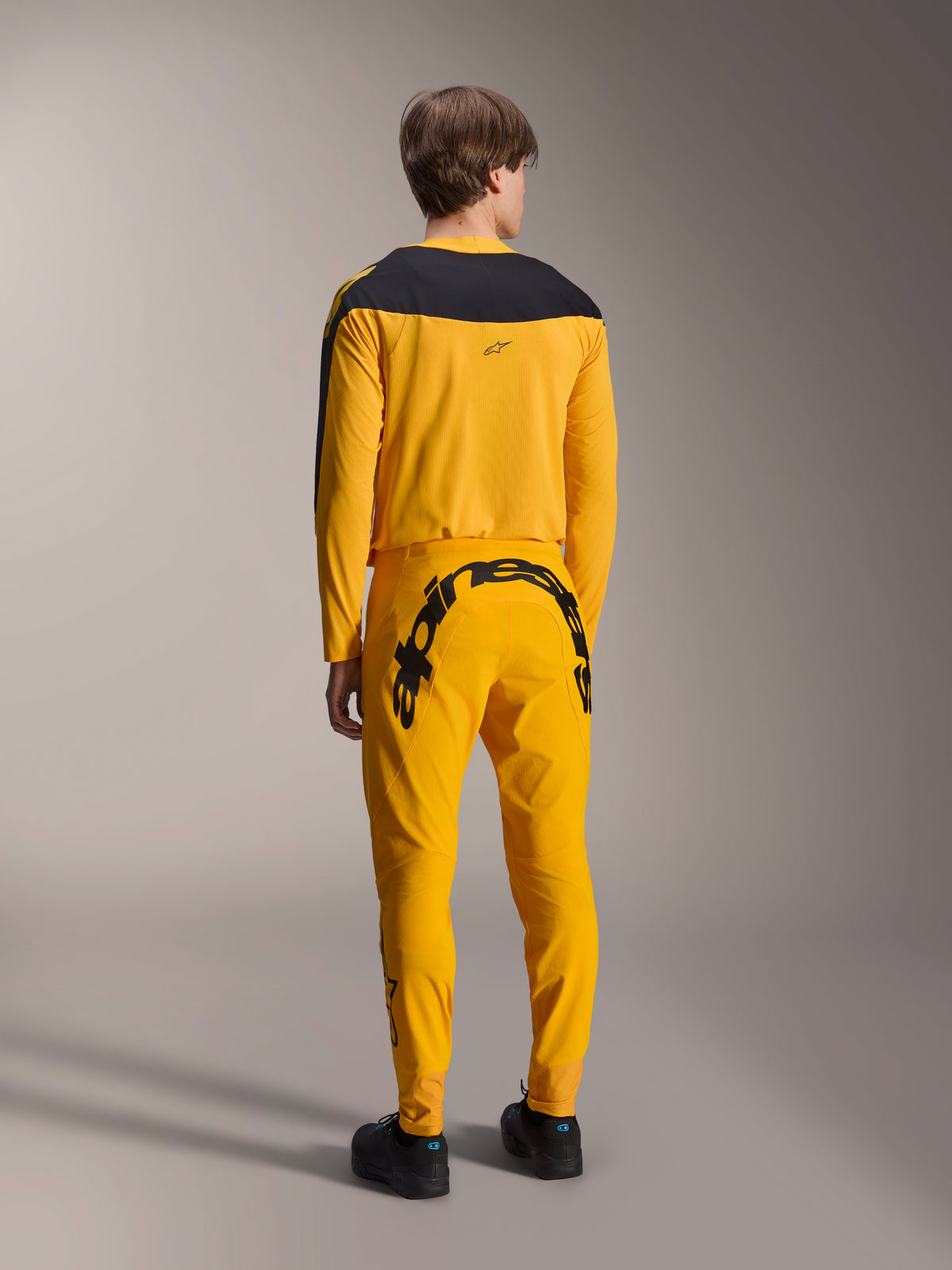 Alpinestars Limited Edition A-Supra Melt Gold gear-set, MTB-jersey en broek, donkergeel en zwart, achteraanzicht met groot Alpinestars-woordmerk op het zitvlak en zwart schouderstuk op de jersey, hoogwaardige mountainbikekleding