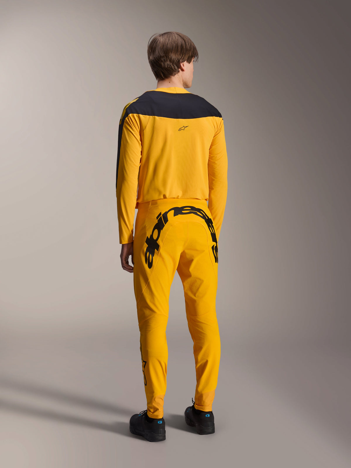 Alpinestars Limited Edition A-Supra Melt Gold Trikot, Langarm-MTB-Trikot, Dunkelgelb mit schwarzem Schultereinsatz, getragen mit passender gelber Hose mit markantem schwarzem Branding auf der Rückseite, Rückansicht eines Performance-Radsport-Kits