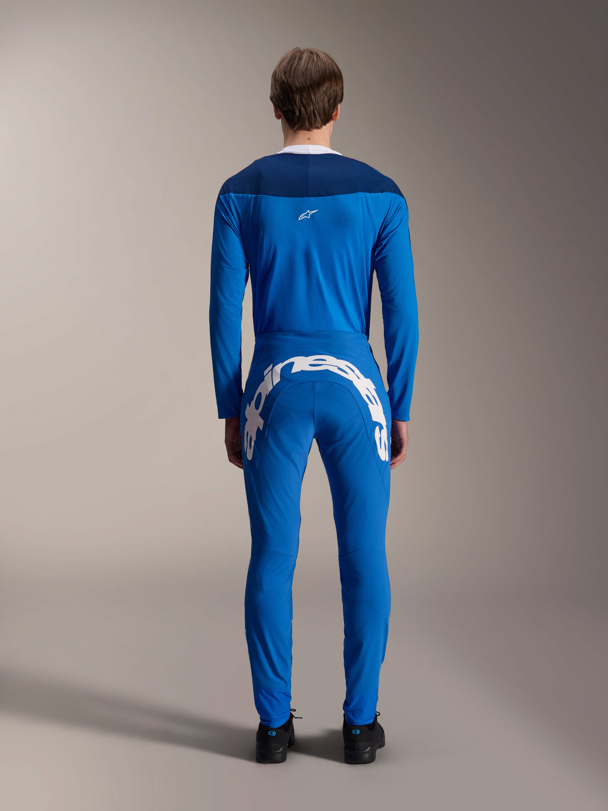 Alpinestars A-Supra Melt broek, blauw, achteraanzicht met een groot wit Alpinestars-woordmerk op de achterpas, gedragen met een bijpassende blauwe jersey met lange mouwen en zwarte schoenen, performance MTB-racekleding