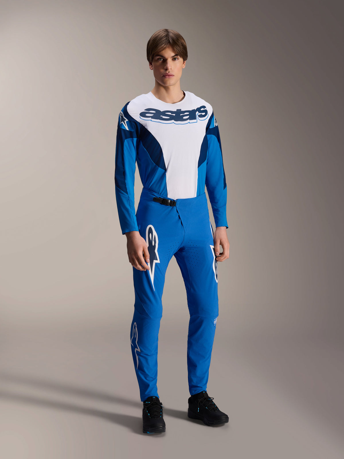 Männliches Model im Alpinestars A-Supra Melt Trikot – Langarm in Blau und Weiß, mit einem auffälligen „astars“-Logo auf der Brust, kombiniert mit einer passenden blauen Mountainbike-Hose und schwarzem Schuhwerk, leistungsorientierte MTB-Rennbekleidung.