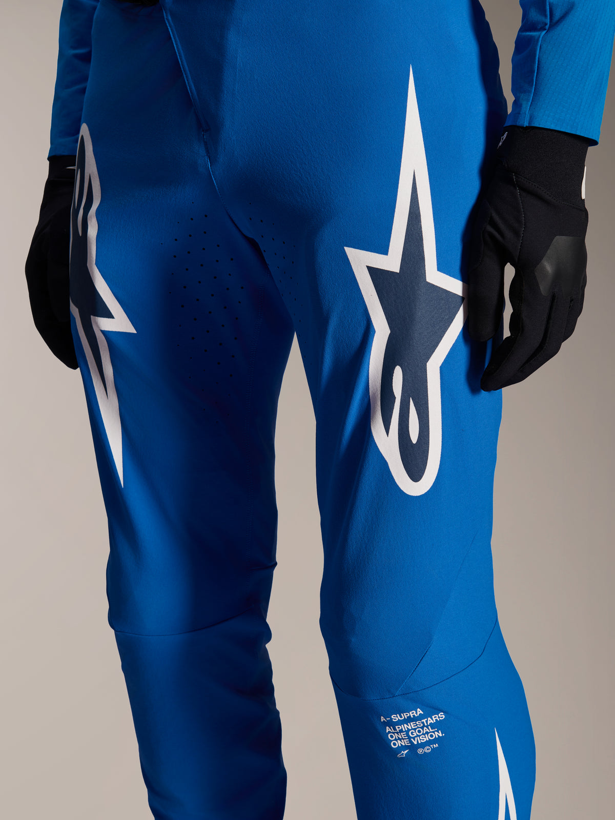 Alpinestars A-Supra Melt broek, blauwe MTB-broek, met grote witte Astars-logo's op de dijen, lasergesneden perforaties voor ventilatie en kenmerkende branding op het onderbeen, gedragen met zwarte handschoenen, performance MTB-kleding