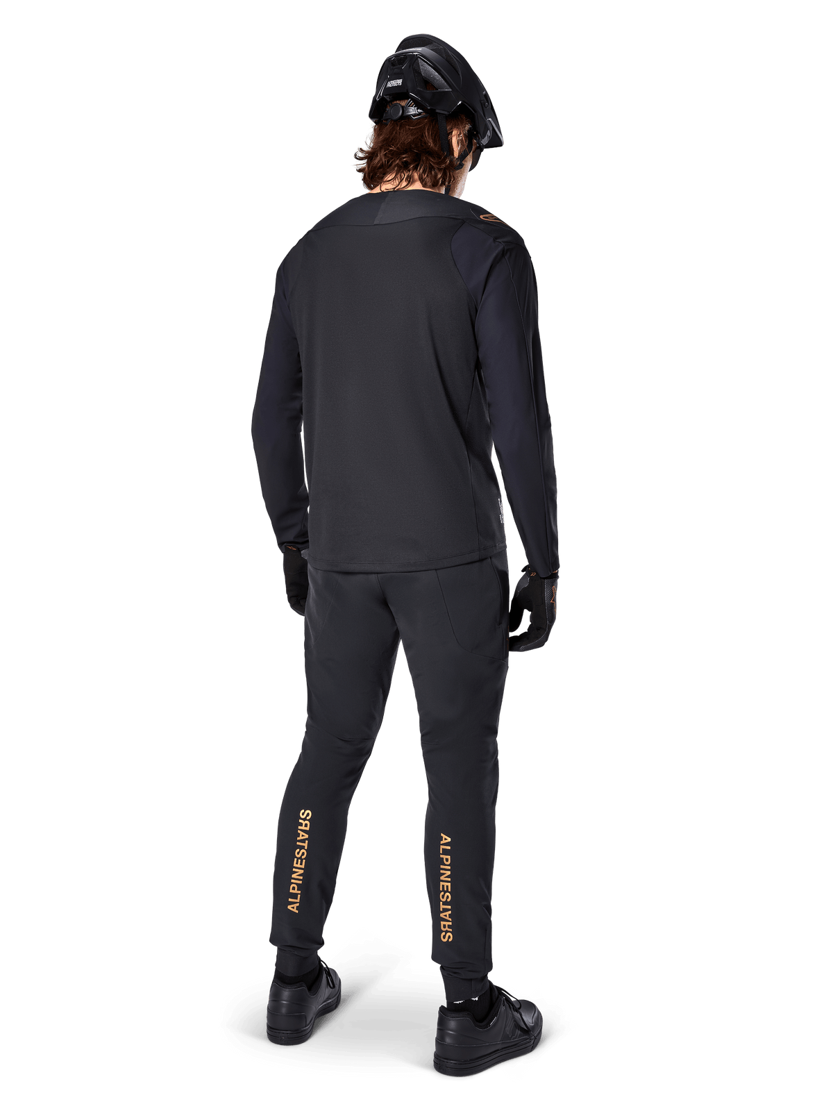 Vue arrière d'un cycliste de VTT portant le maillot à manches longues Alpinestars A-Aria Polartec Switch noir, associé à un pantalon de VTT noir avec logo Alpinestars beige sur les mollets, casque et gants noirs, équipement de cyclisme professionnel pour le tout-terrain.
