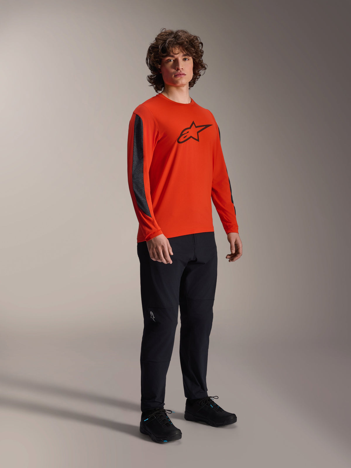 Male model wearing Alpinestars A-Dura Insert Astar Jersey, Long Sleeve, Spicy Orange with black Astar logo on the chest, dark grey sleeve inserts, worn with black pants, professional MTB cycling apparel