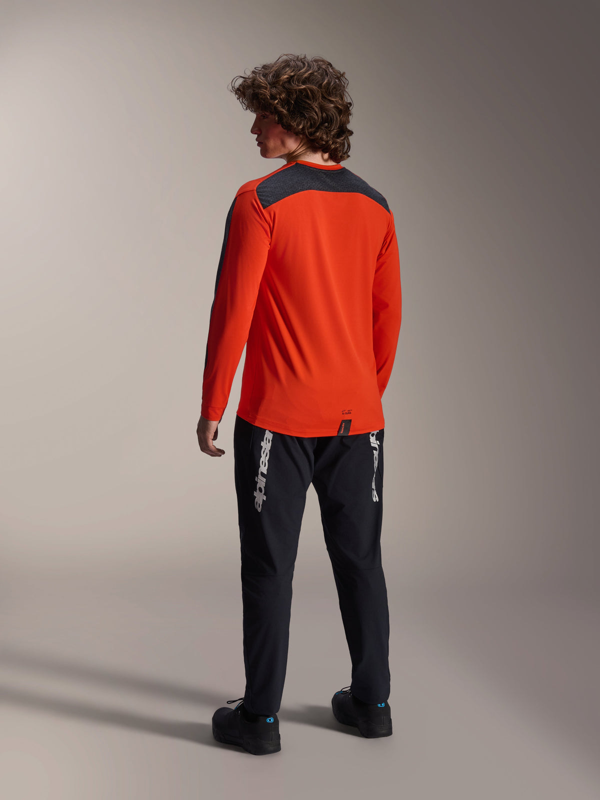 Rear view of a model wearing the Alpinestars A-Dura Insert Astar Jersey in Spicy Orange, long sleeve MTB jersey with dark gray shoulder panel, paired with black branded pants, off-road cycling gear.