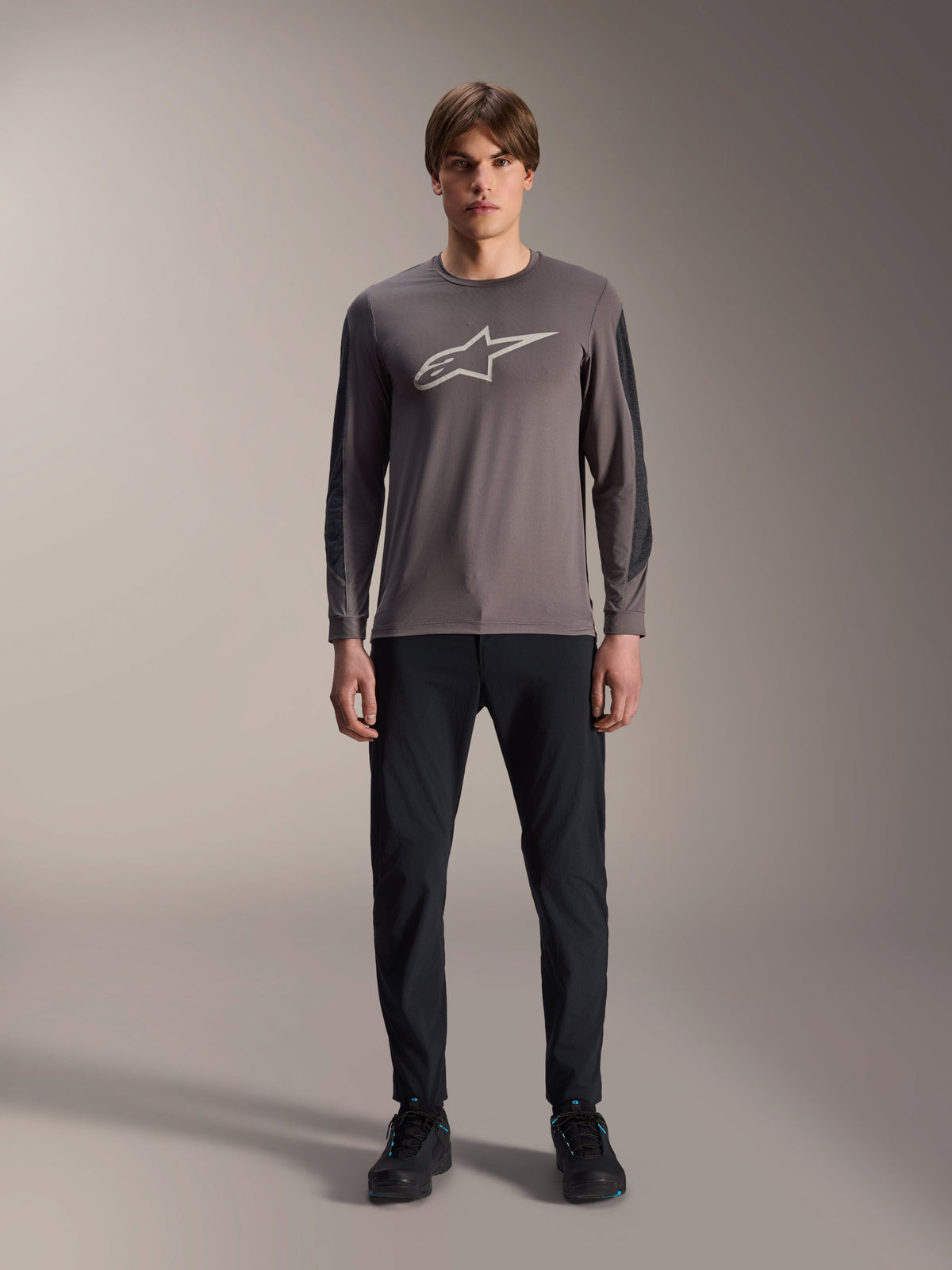 Alpinestars A-Dura Insert Astar Jersey, MTB-shirt met lange mouwen, donkergrijs, met een opvallend lichtgrijs Astar-logo op de borst en donkerdere mouwinzetstukken met textuur, gedragen door een model met een zwarte broek en schoenen, performance kleding voor mountainbiken