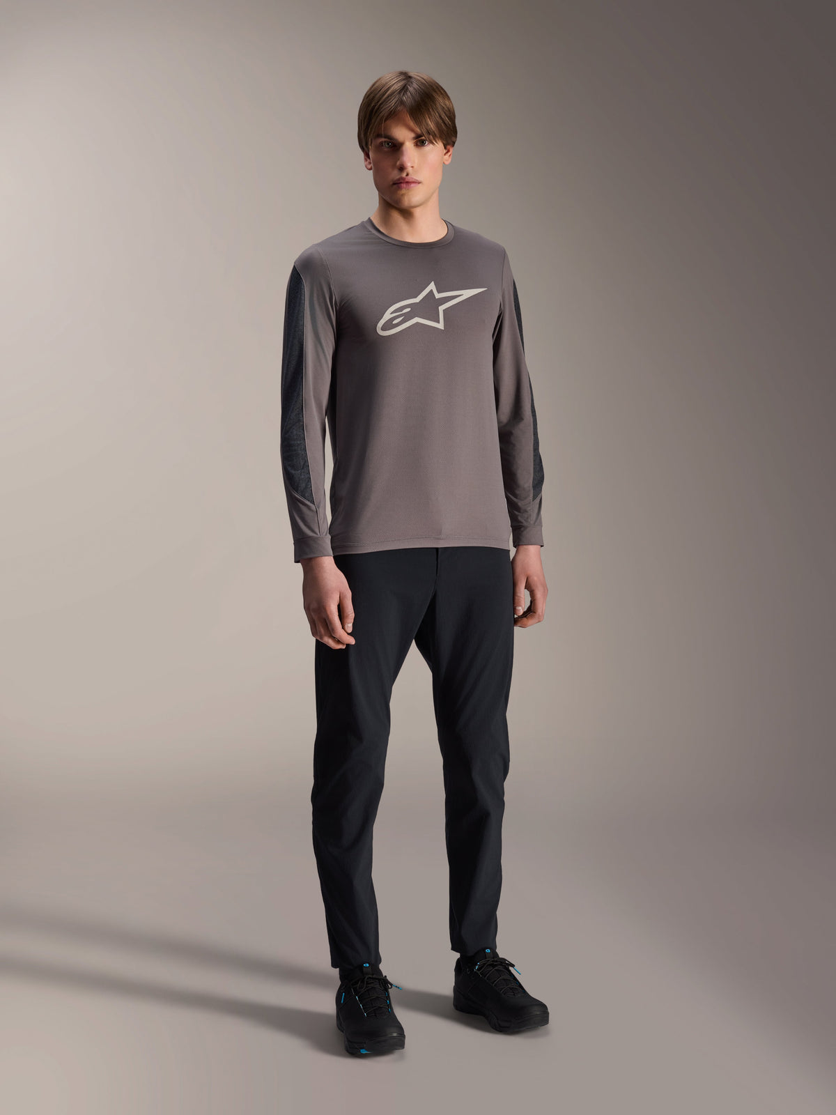 Alpinestars A-Dura Insert Astar Jersey, MTB-shirt met lange mouwen, donkergrijs, met groot wit logo op de borst, gedragen met zwarte technische broek en zwarte schoenen, off-road fietskleding