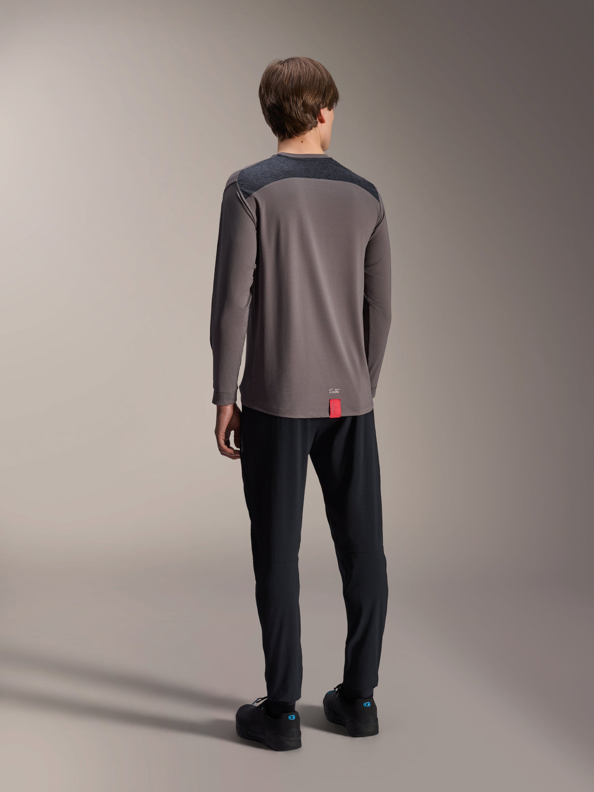 Alpinestars A-Dura Insert Astar Jersey met lange mouwen, donkergrijs, achteraanzicht op model, voorzien van antracietkleurige versterkte schouderpanelen en een klein rood label aan de zoom, gecombineerd met een zwarte broek en schoenen, technische MTB-kleding