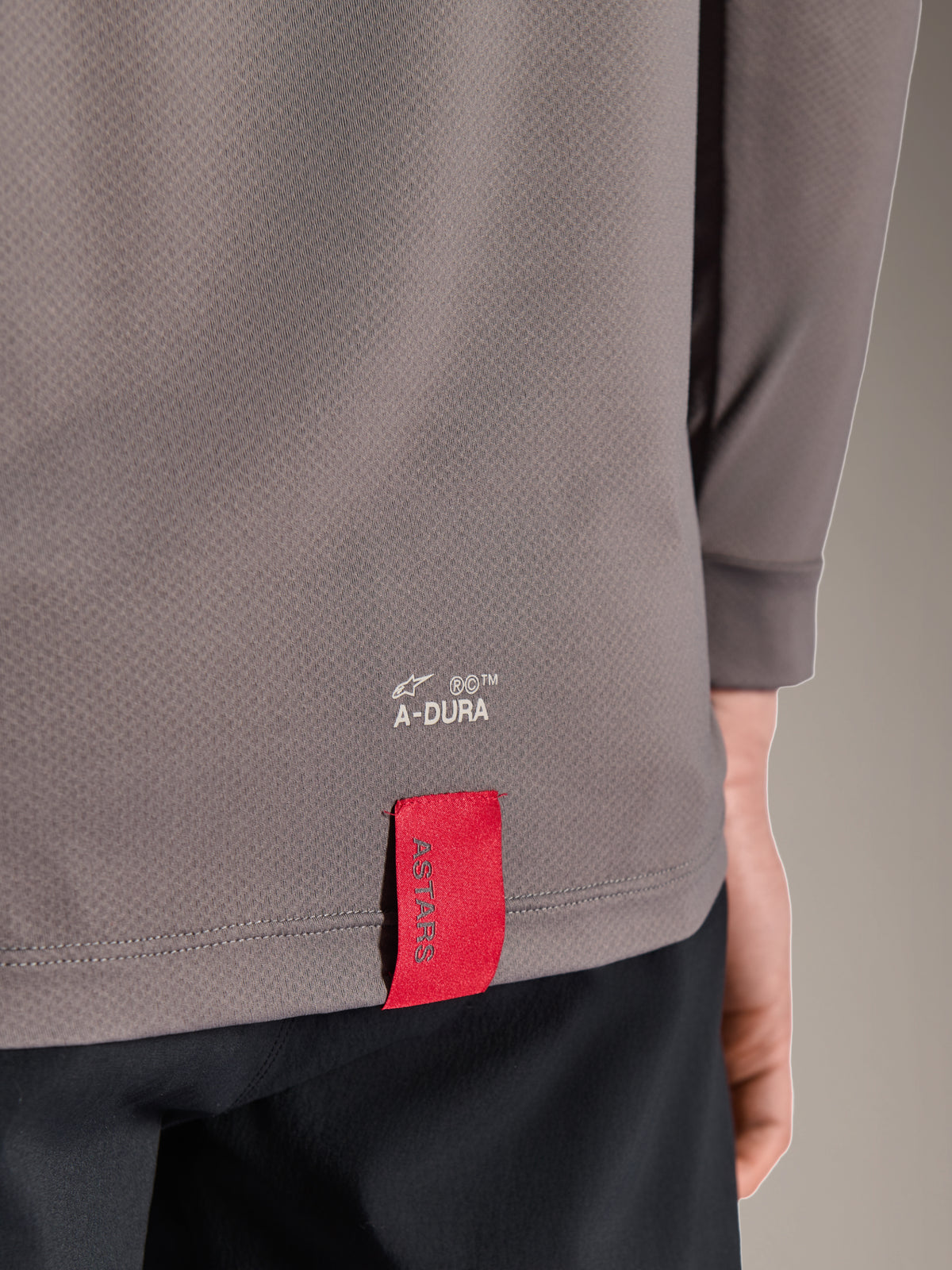 Alpinestars A-Dura Insert Astar Jersey, MTB-jersey met lange mouwen, donkergrijs, close-up detail van de zoom onderaan de rug met witte A-DURA-branding en een kenmerkend rood ASTARS-stoffen label, getextureerde technische stof